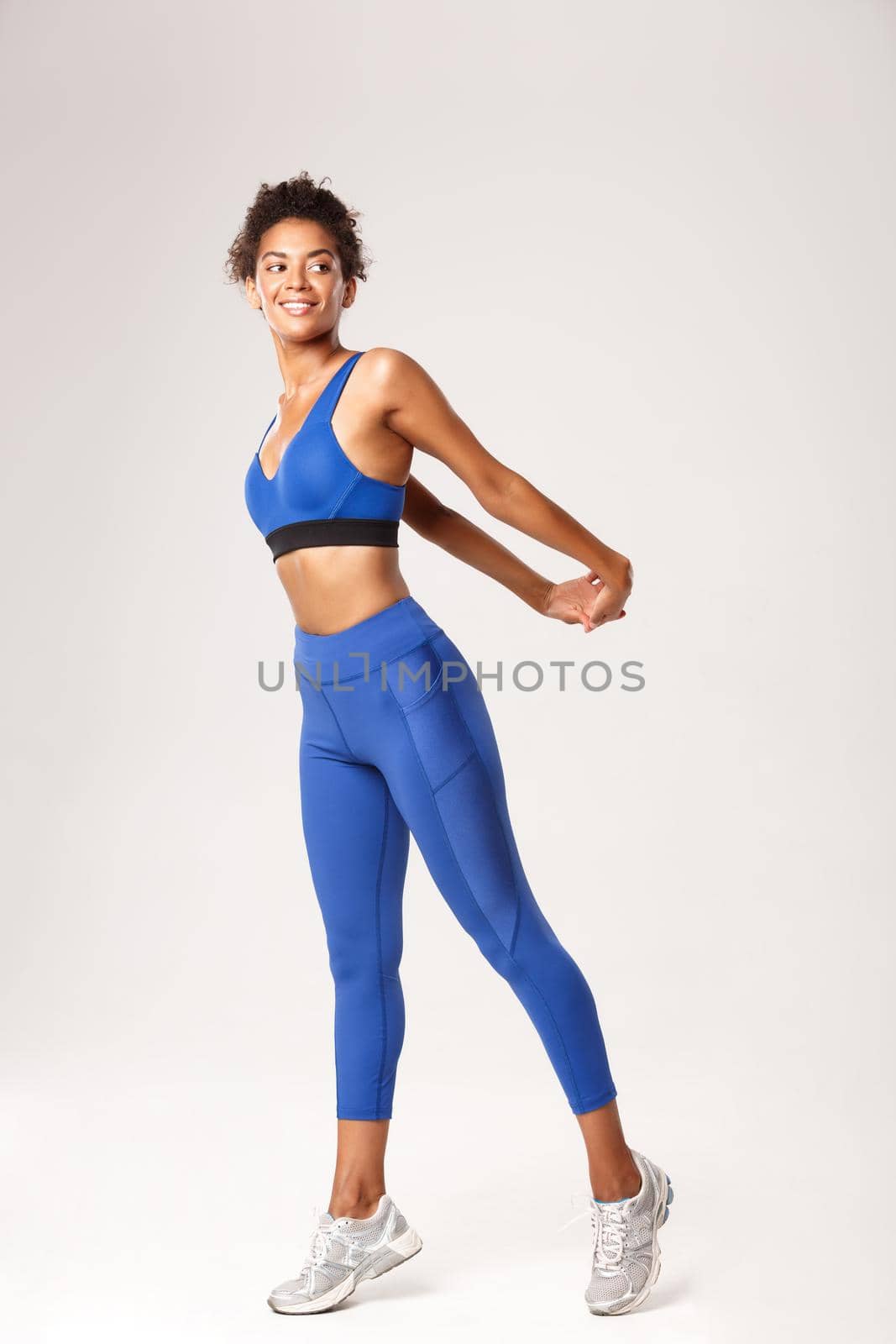 Full length of smiling african-american sportswoman in blue sport outfit, stretching before workout, looking right, standing over white background.