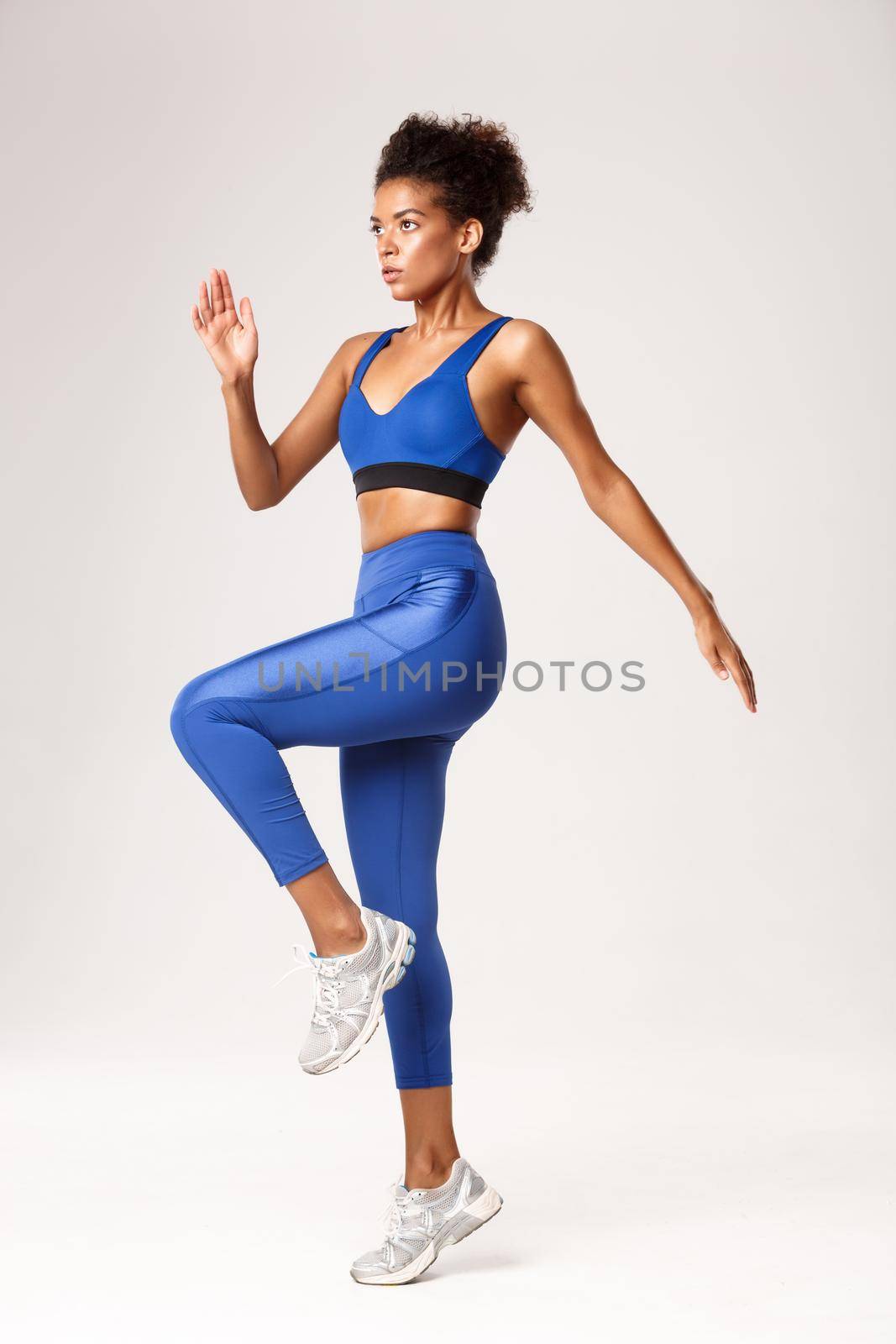 Full length profile shot of african-american sportswoman in fitness clothing, lift leg and looking serious during workout, exercising over white background.