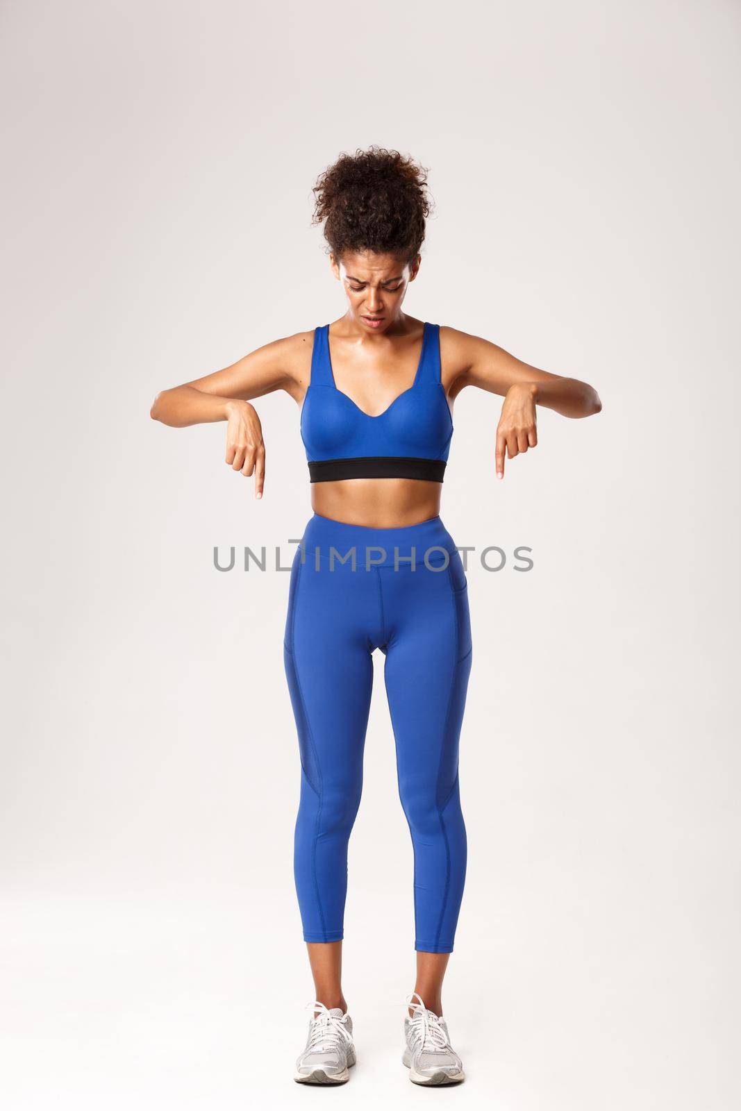 Full length of upset african-american fitness girl in blue sportswear, looking and pointing fingers down, frowning with worried and sad expression, standing over white background.