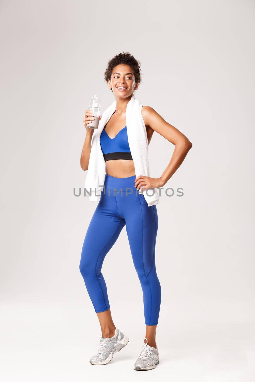 Full length of smiling fit and healthy african-american female athlete workout and drinking water, standing with towel against white background by Benzoix