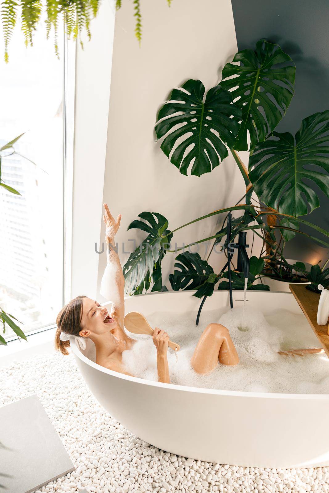 Cute young woman singing into a brush while bathing in the bathtub decorated with tropical plant. Wellness, body care concept