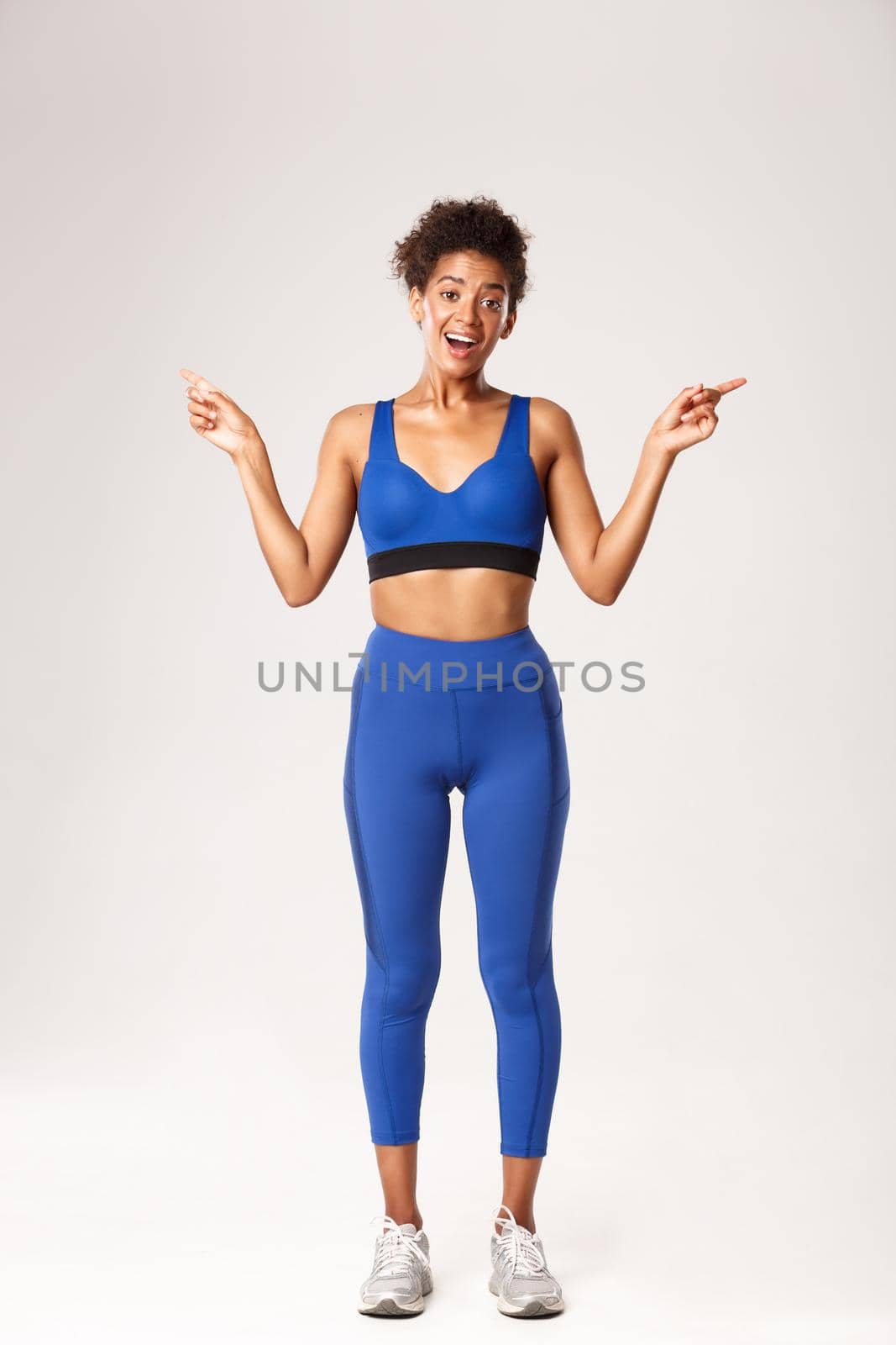 Full length of excited and happy african-american fitness girl, wearing blue sport outfit, pointing fingers sideways, showing left and right logo with pleased face, white background by Benzoix