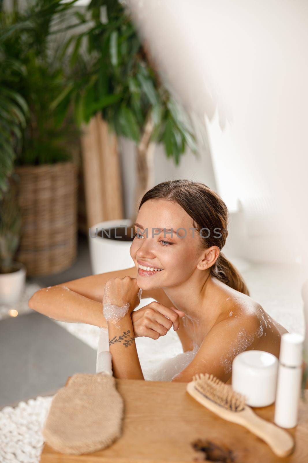 Joyful young woman smiling away and leaning on bathtub side while bathing at spa resort. Wellness, beauty and care concept