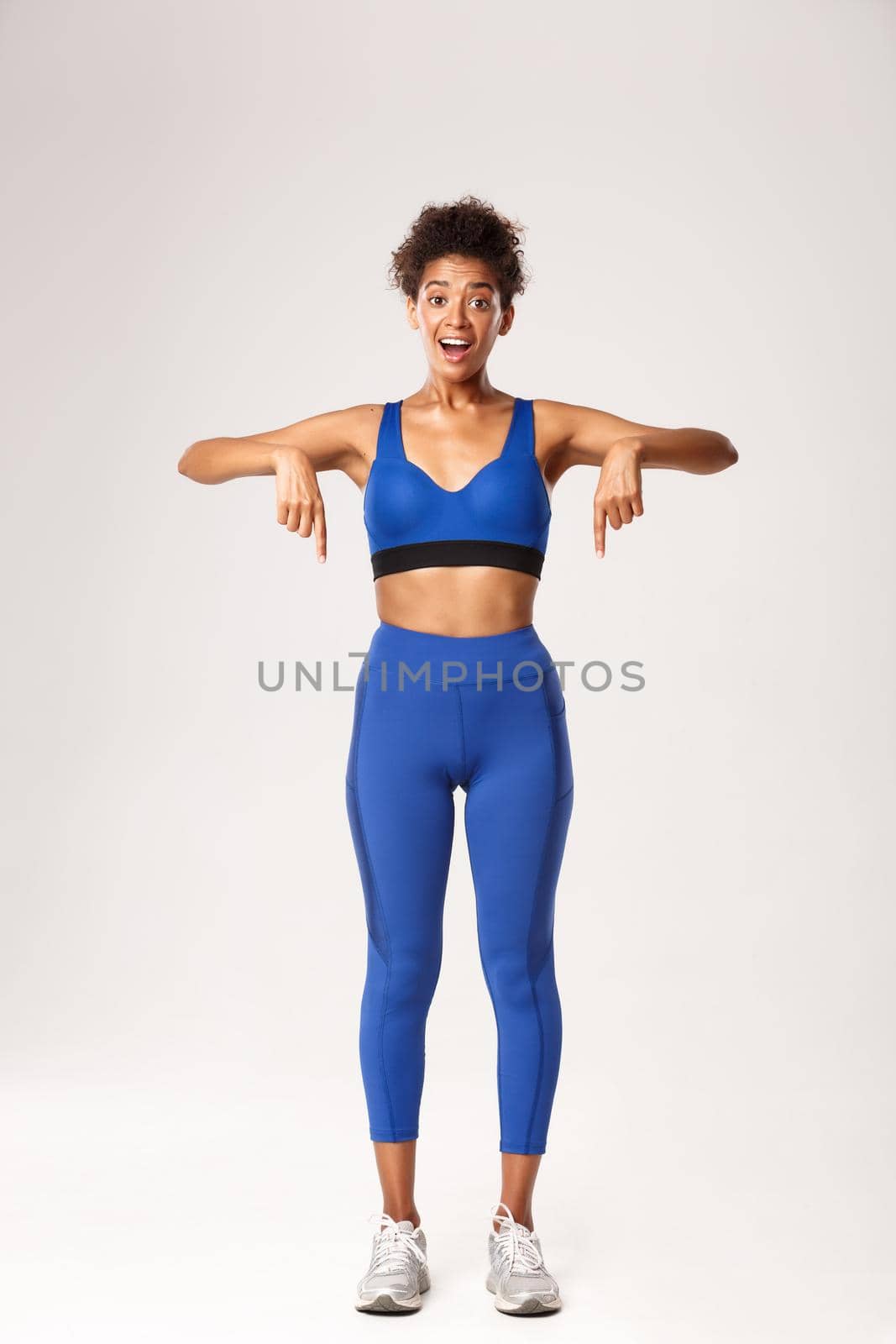 Full length of surprised african-american fitness girl, wearing blue sports outfit, pointing fingers down with amused smile, standing against white background.