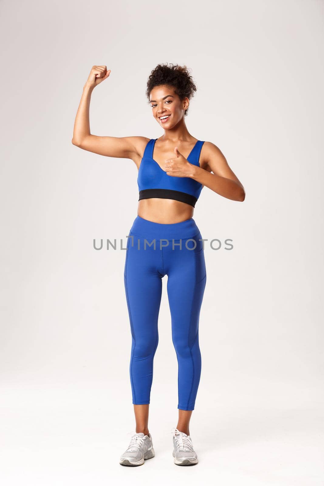 Full length of strong and attractive african american sportswoman in blue sportswear, showing thumbs-up and flex biceps, recommend gym workout, good training session, white background.