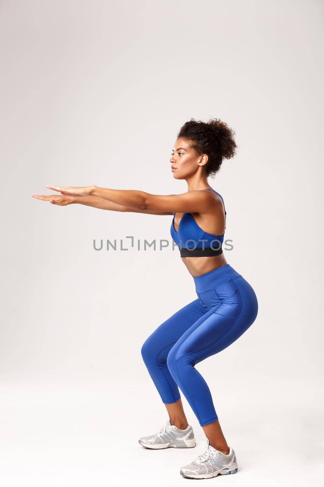 Full length profile shot of determined african-american fitness woman, wearing blue sport outfit, squat with hands stretched foward, workout over white background.