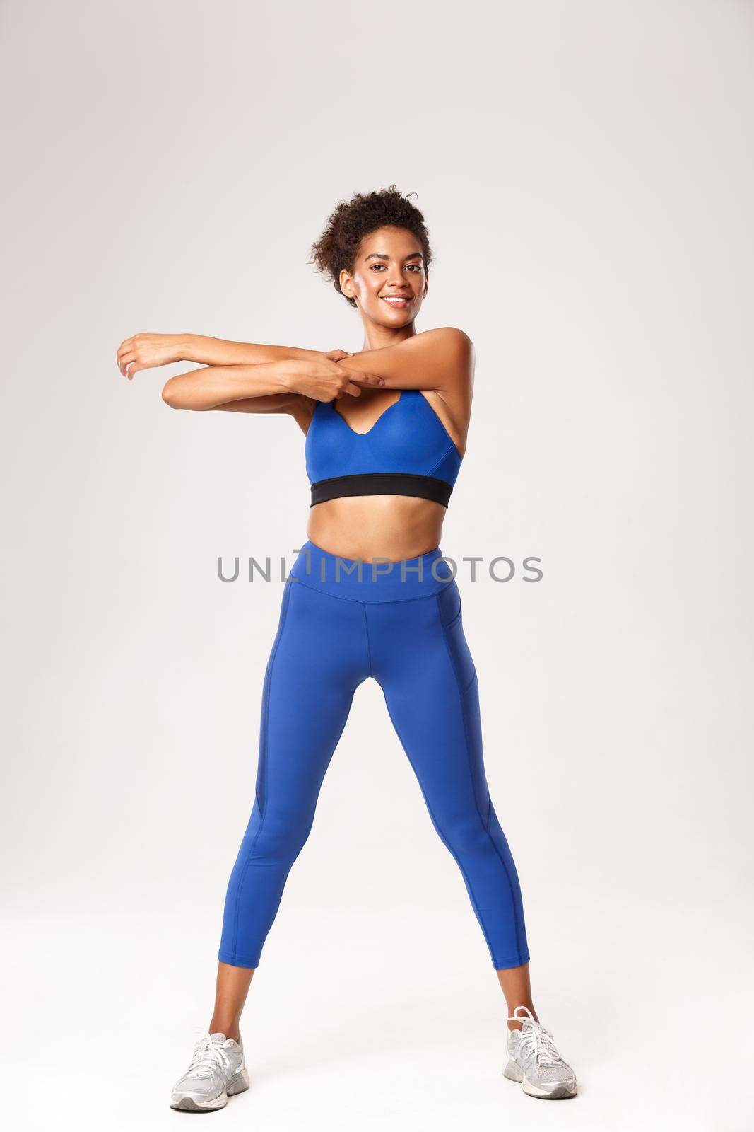 Full length of smiling athletic african-american sportswoman, wearing blue sport outfit, stretching before fitness activity, workout over white background.