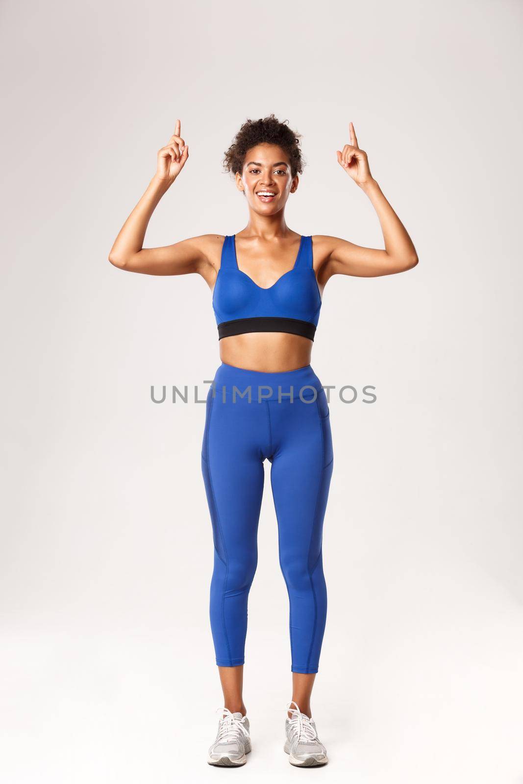Full length of attractive slim african-american sportswoman, athlete pointing fingers up at logo or brand advertisement, standing in workout outfit over white background.