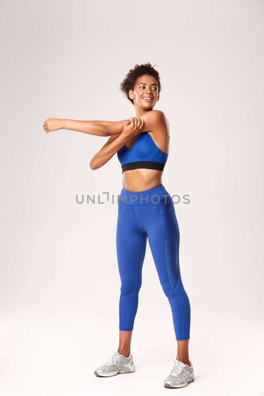 Full length of attractive african-american sportswoman, looking excited before workout, stretching hands and smiling, standing over white background by Benzoix