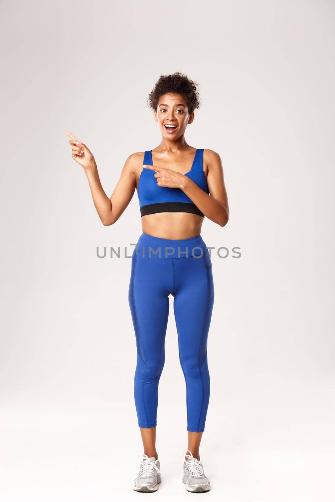 Full length of good-looking african-american sportswoman in blue sportswear, pointing fingers left, showing advertisement about sport and workout, standing over white background.