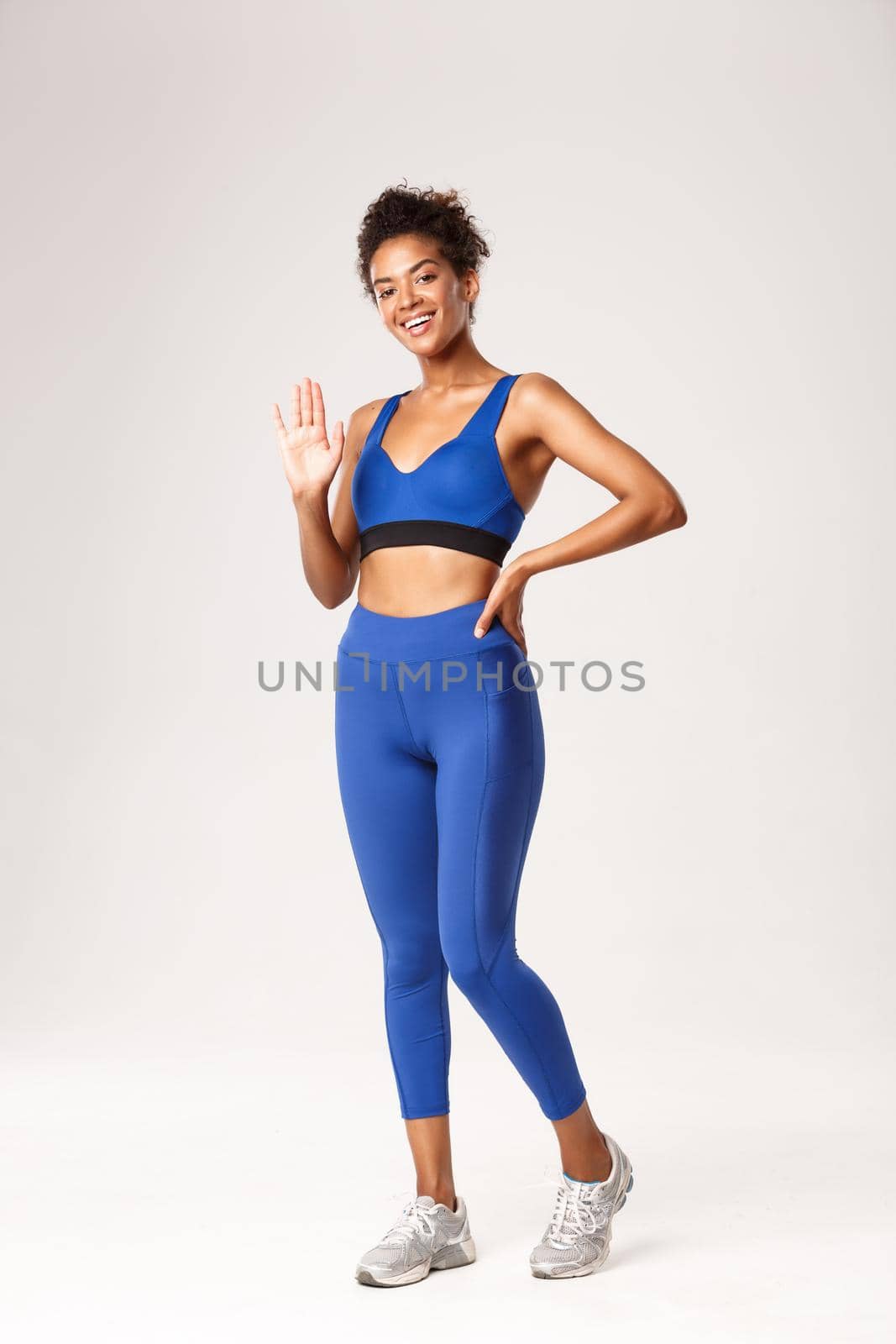 Full length of smiling girl in gym clothing saying hello, standing over white background in sportswear and waving hand, greeting someone, standing over white background by Benzoix