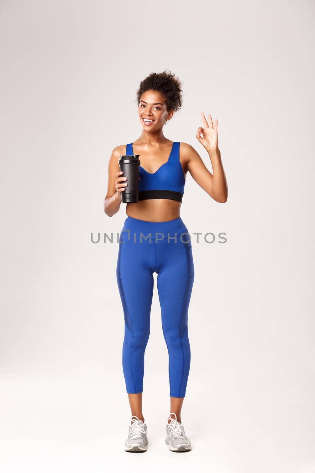 Full length of smiling african-american fitness woman looking healthy and fit, showing okay sign, drinking water during workout, standing against white background.