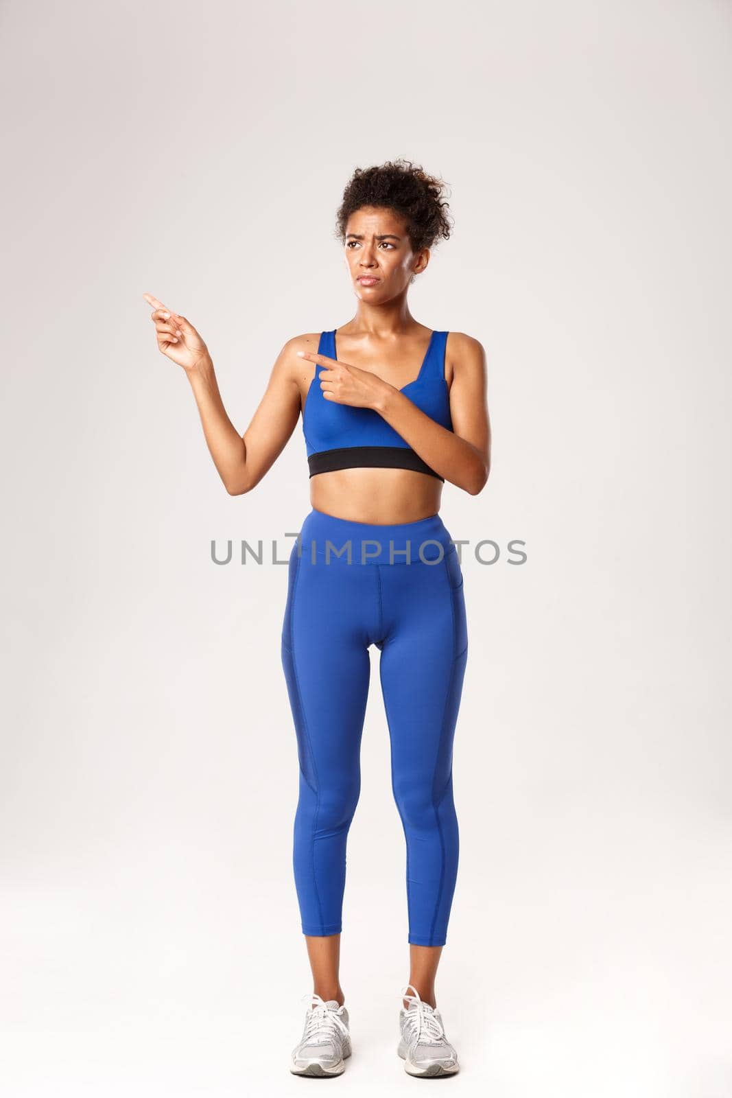 Full length of attractive african-american sportswoman looking upset, pointing fingers left and sulking, express disappointment or dislike, standing in blue sportswear, white background.