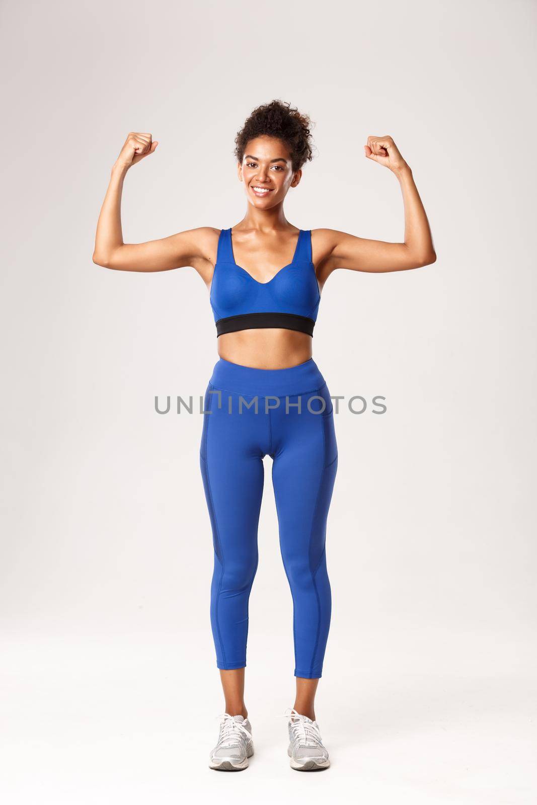 Full length of strong and confident african american girl showing her muscles, flex biceps and smiling, demonstrate gym and workout progress, standing voer white background.