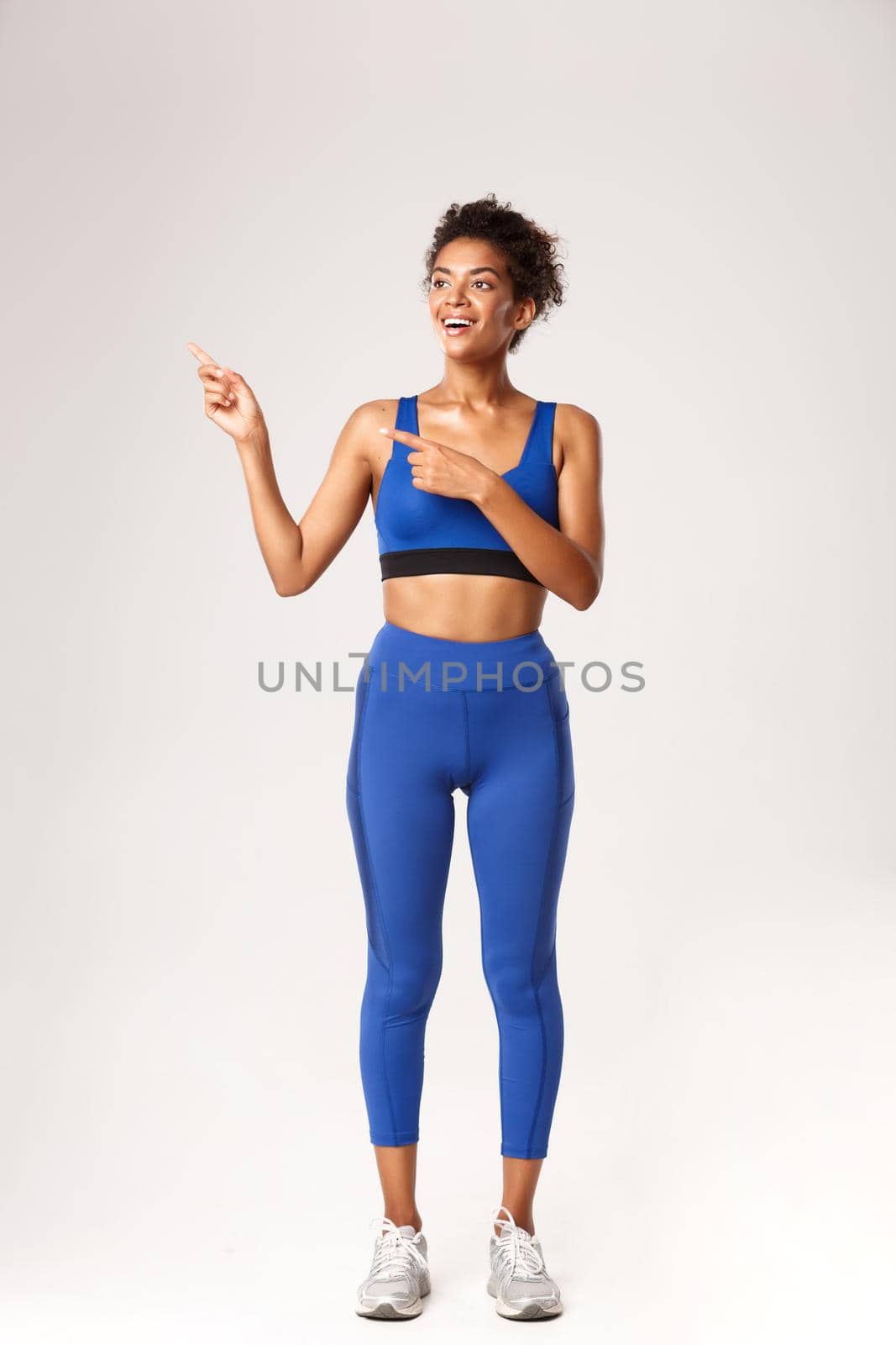 Full length of surprised smiling african-american sportswoman, looking happy at something, pointing fingers left, standing over white background.