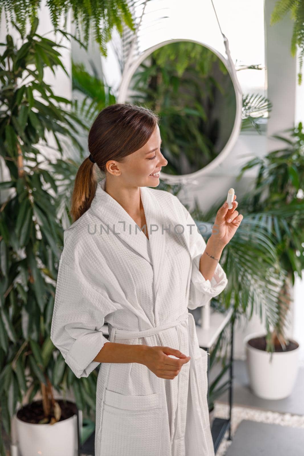 Lovely young woman in white bathrobe smiling while holding lip balm, standing in the bathroom decorated with plants. Skincare cosmetics concept