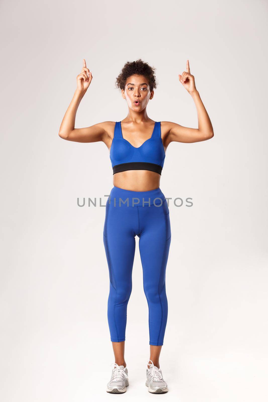 Full length of amazed good-looking fitness girl in blue sports bra and leggings, pointing fingers up, showing promo about workout or gym, standing over white background.