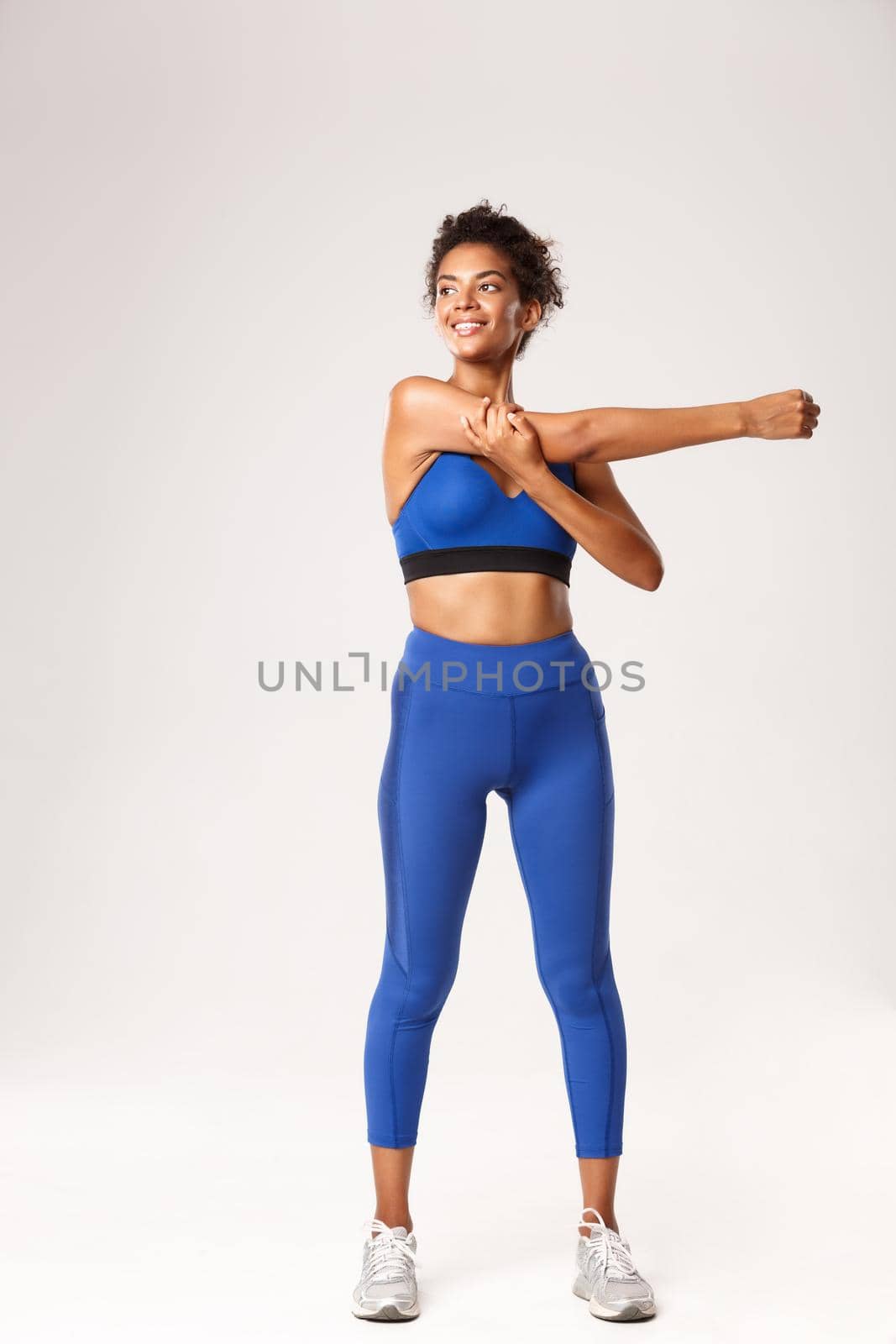 Full length of smiling beautiful african-american sportswoman, stretching hands, warming-up before workout and looking left determined, standing over white background.