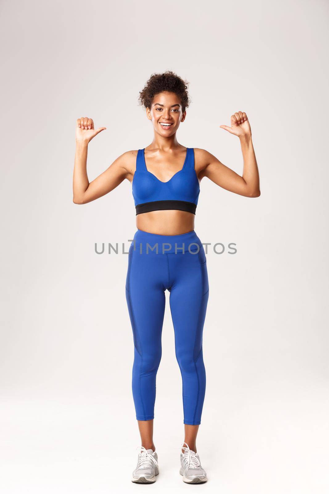 Full length of happy confident african-american sportswoman in blue sport outfit, pointing at herself with proud cheerful smile, showing workout progress, standing over white background.
