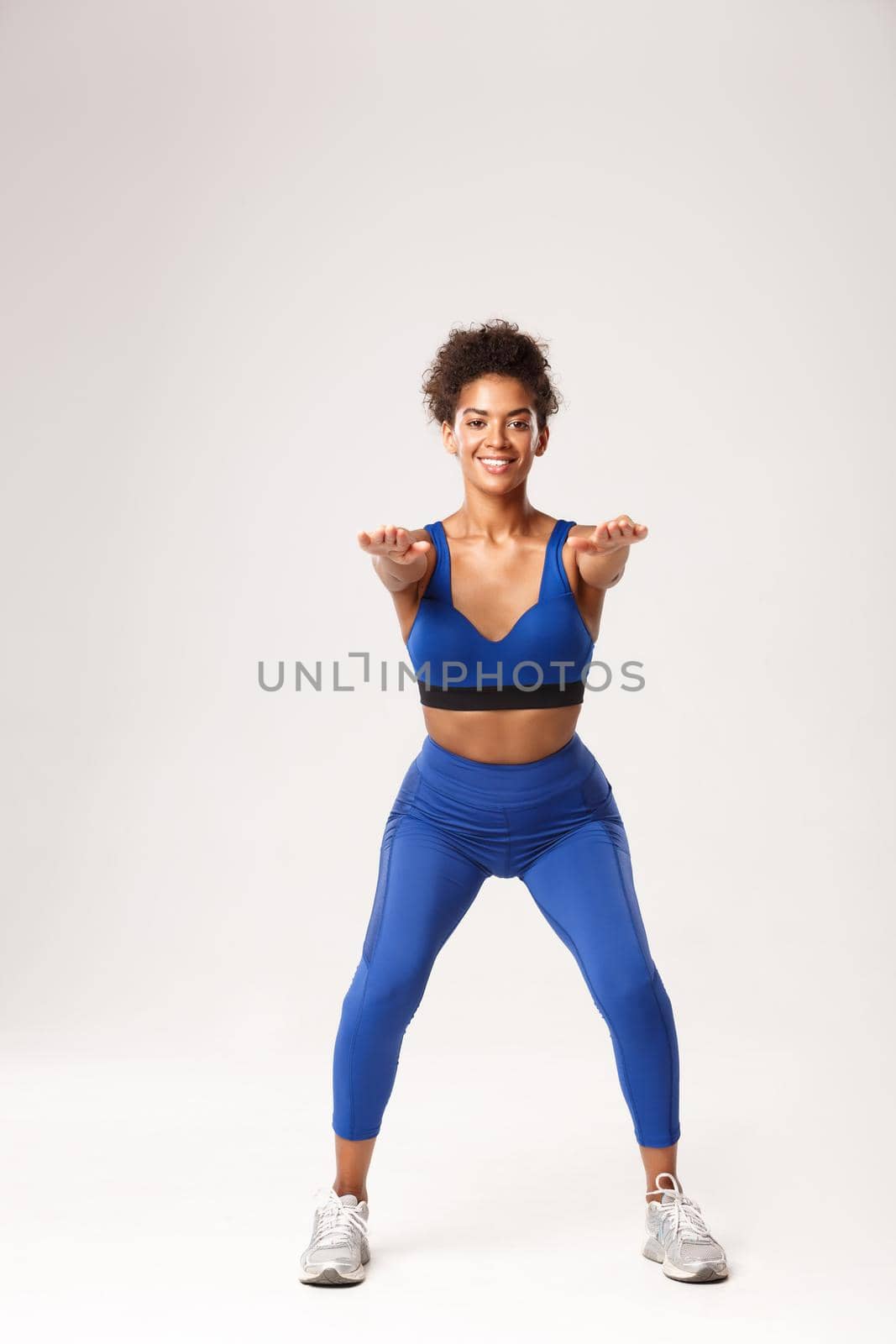 Full length of attractive african-american fit woman in sport clothing, stretch hands foward and making squats exercises, smiling at camera, standing against white background by Benzoix