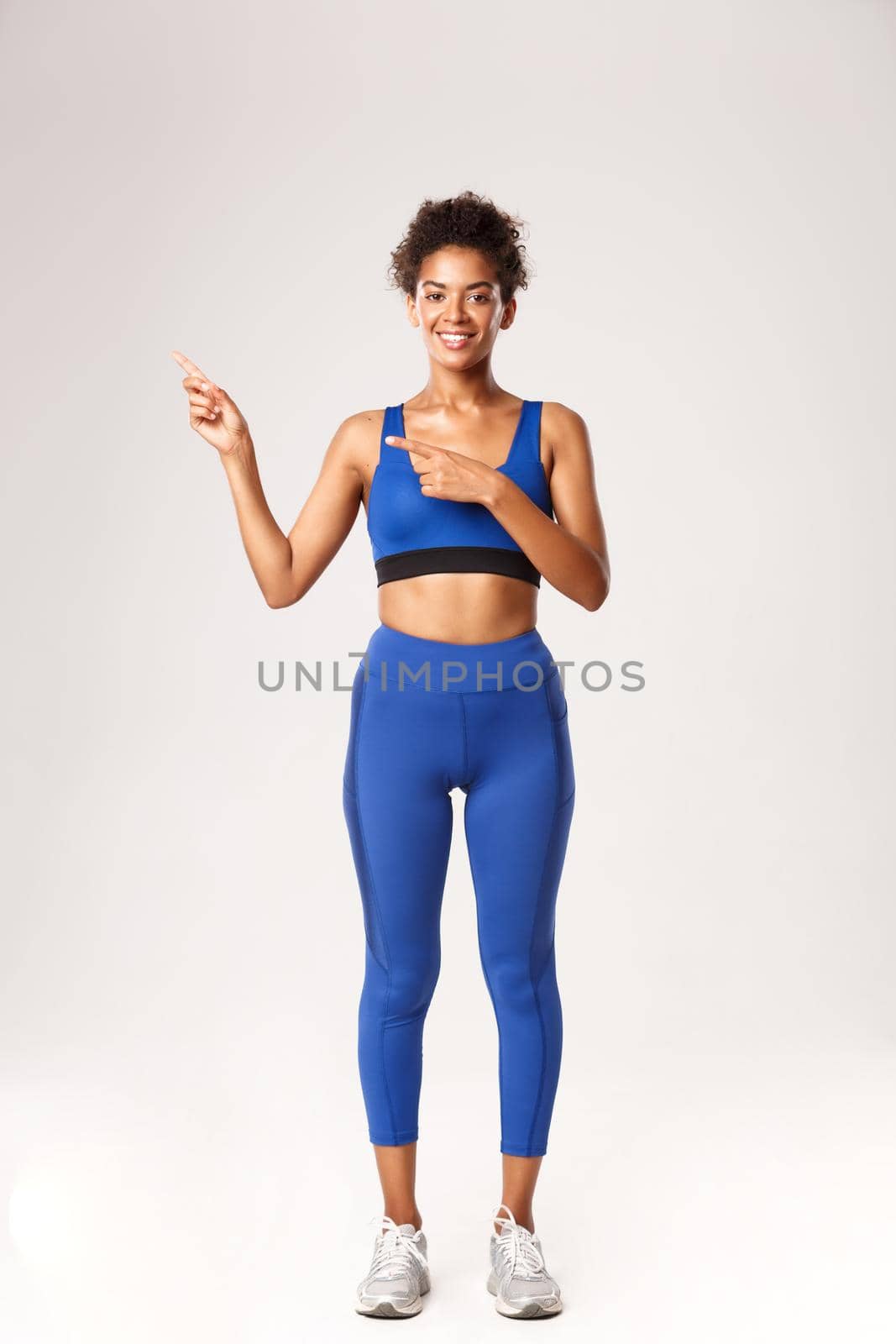 Full length of smiling attractive african-american girl in fitness clothing, pointing fingers left, showing gym logo or product banner, standing over white background.