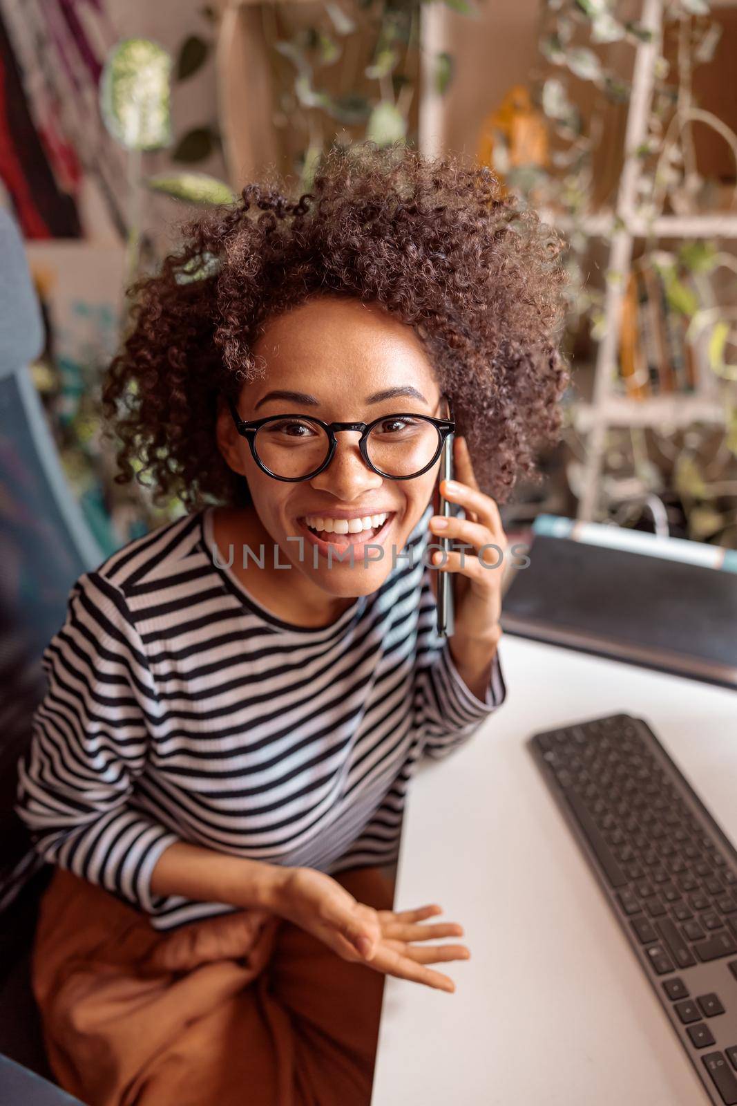 Cheerful multiethnic lady having phone conversation and smiling while sitting at the table with keyboard