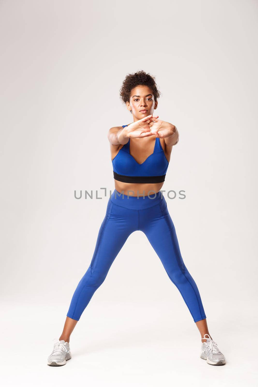 Full length of focused fitness girl stretching and warming-up before workout. African-american gym coach showing exercises, standing over white background.