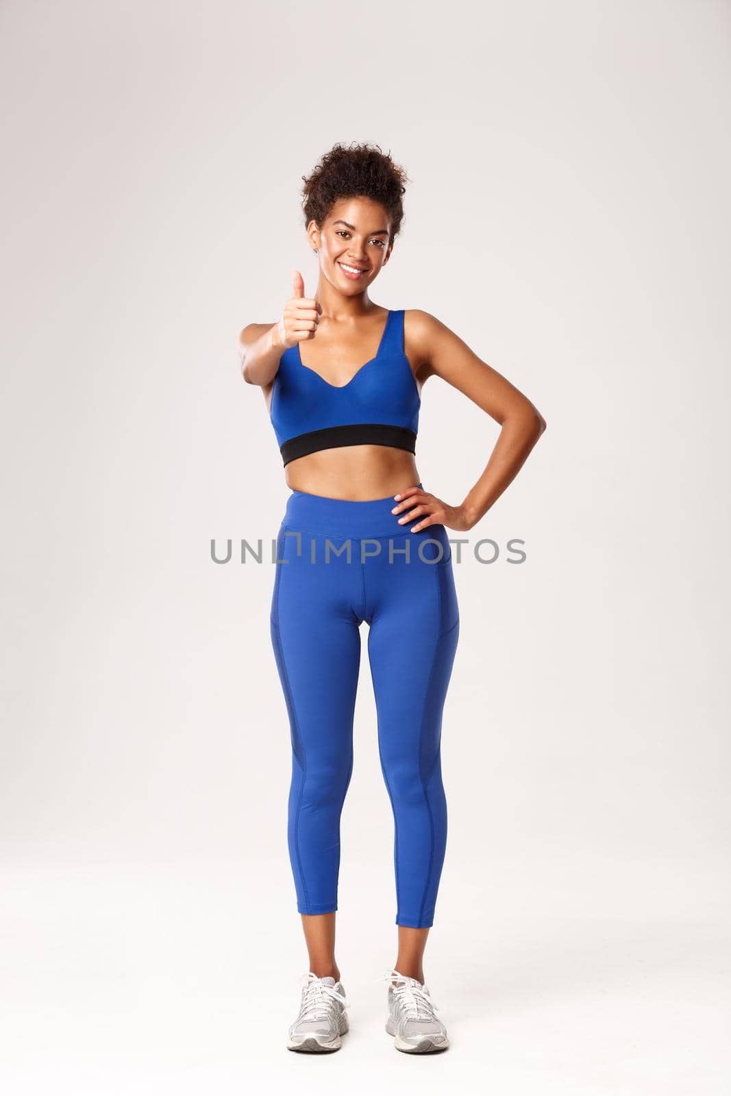Full length of attractive african american sportswoman in fitness clothing, showing thumbs-up in approval, recommend sport brand, standing over white background.
