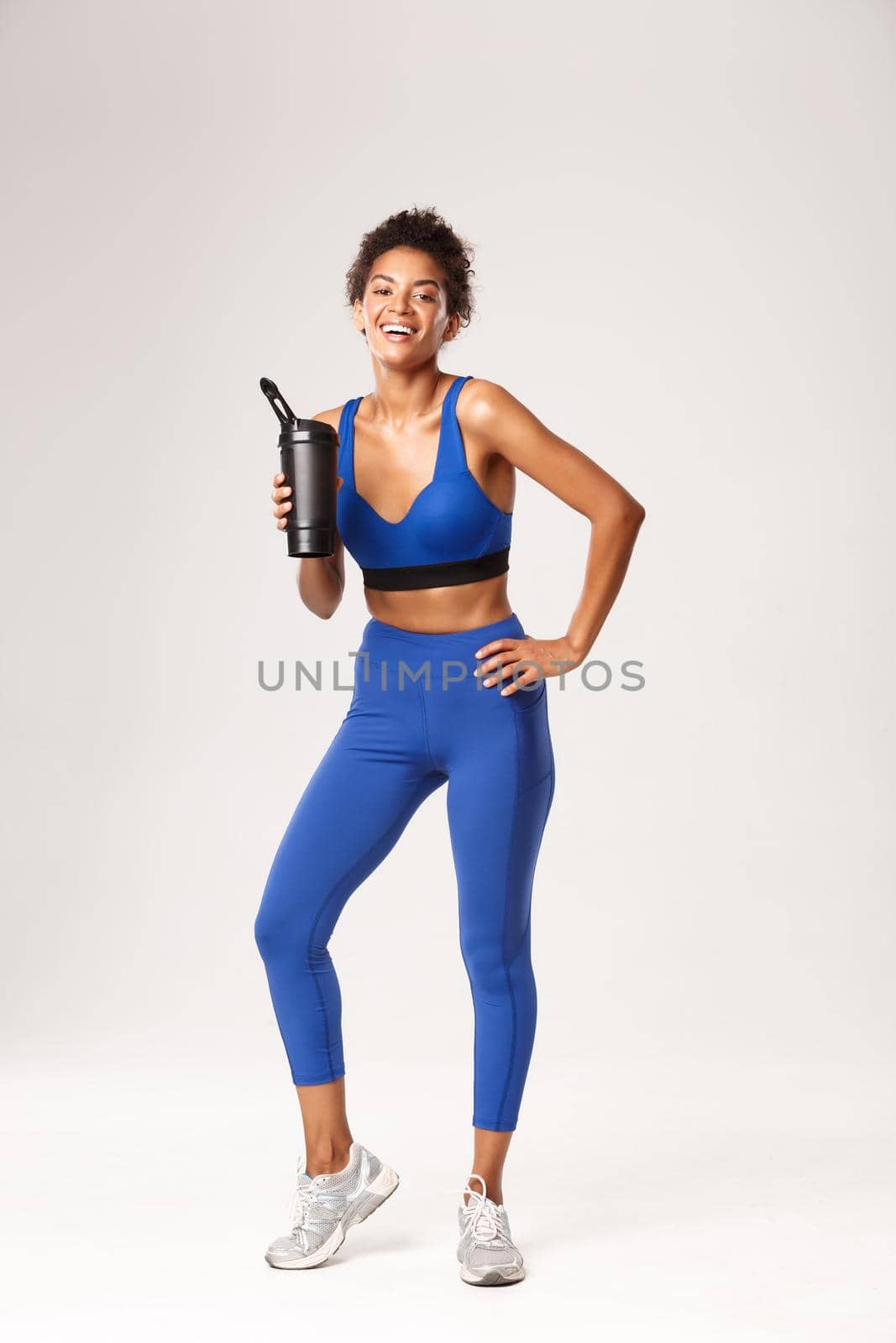 Full length of attractive healthy fitness woman in blue sport uniform, drinking water or protein from bottle, laughing and smiling after good workout, standing against white background by Benzoix