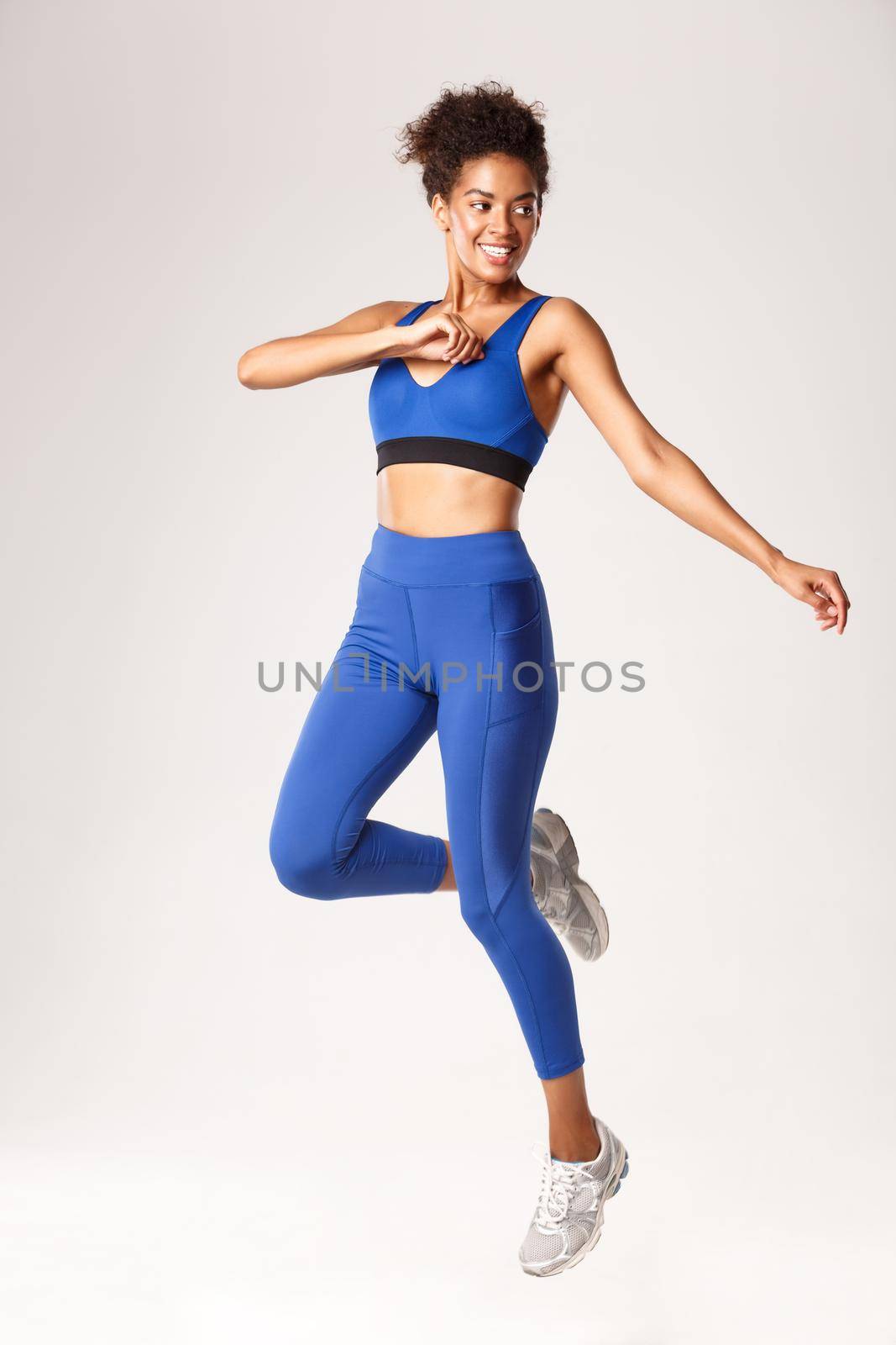 Full length of smiling attractive african-american sportswoman, wearing blue sportswear, jumping and looking away with determined face, worout over white background.