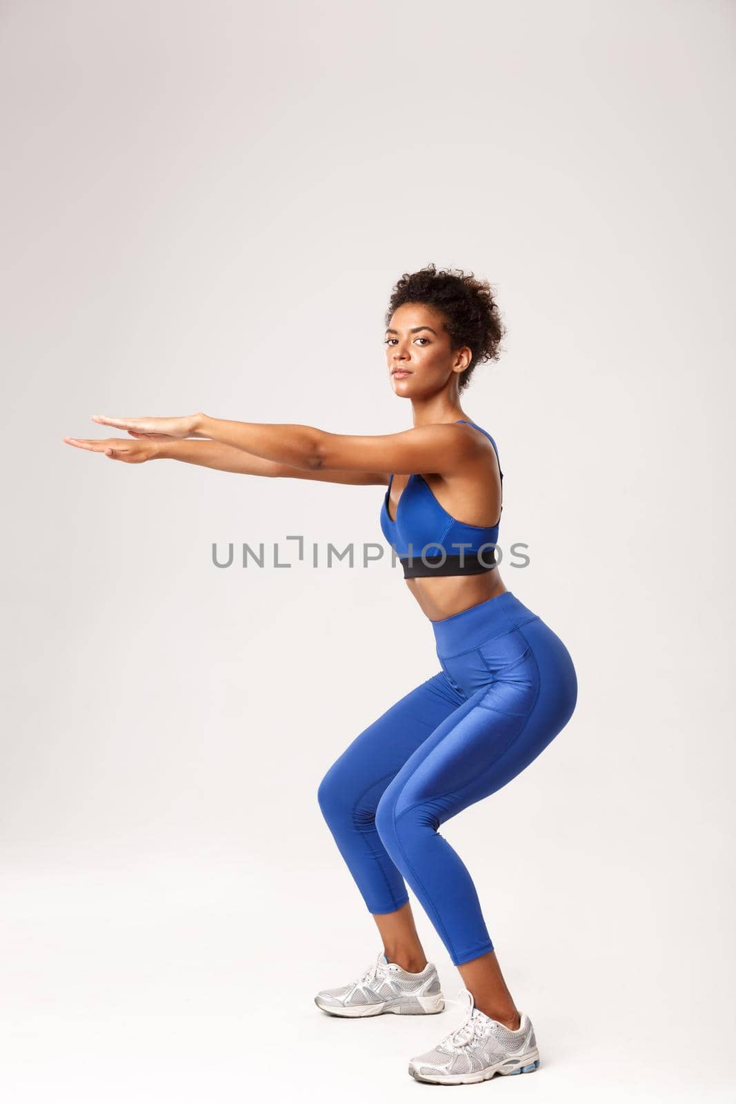 Full length of beautiful and healthy african-american woman in blue sport costume, stretching hands for squats exercises, looking at camera, worout over white background.