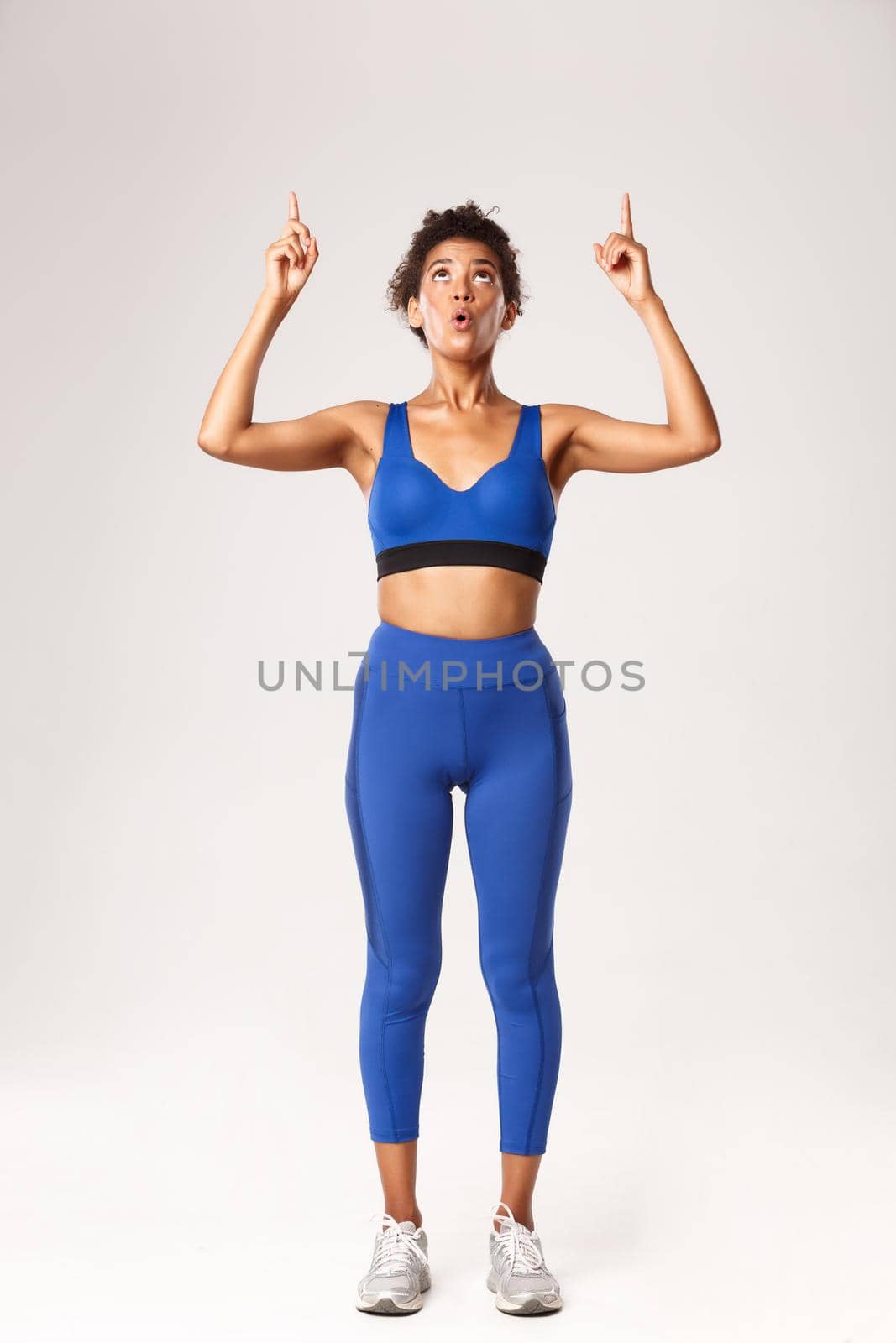 Full length of amazed african-american sportswoman, looking and pointing fingers up with wondered face, standing in blue sports costume over white background by Benzoix