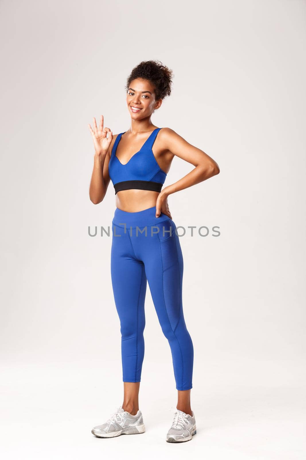 Full length of african american sportswoman with athletic body, showing okay gesture in approval, recommend gym or workout plan, standing over white background satisfied.