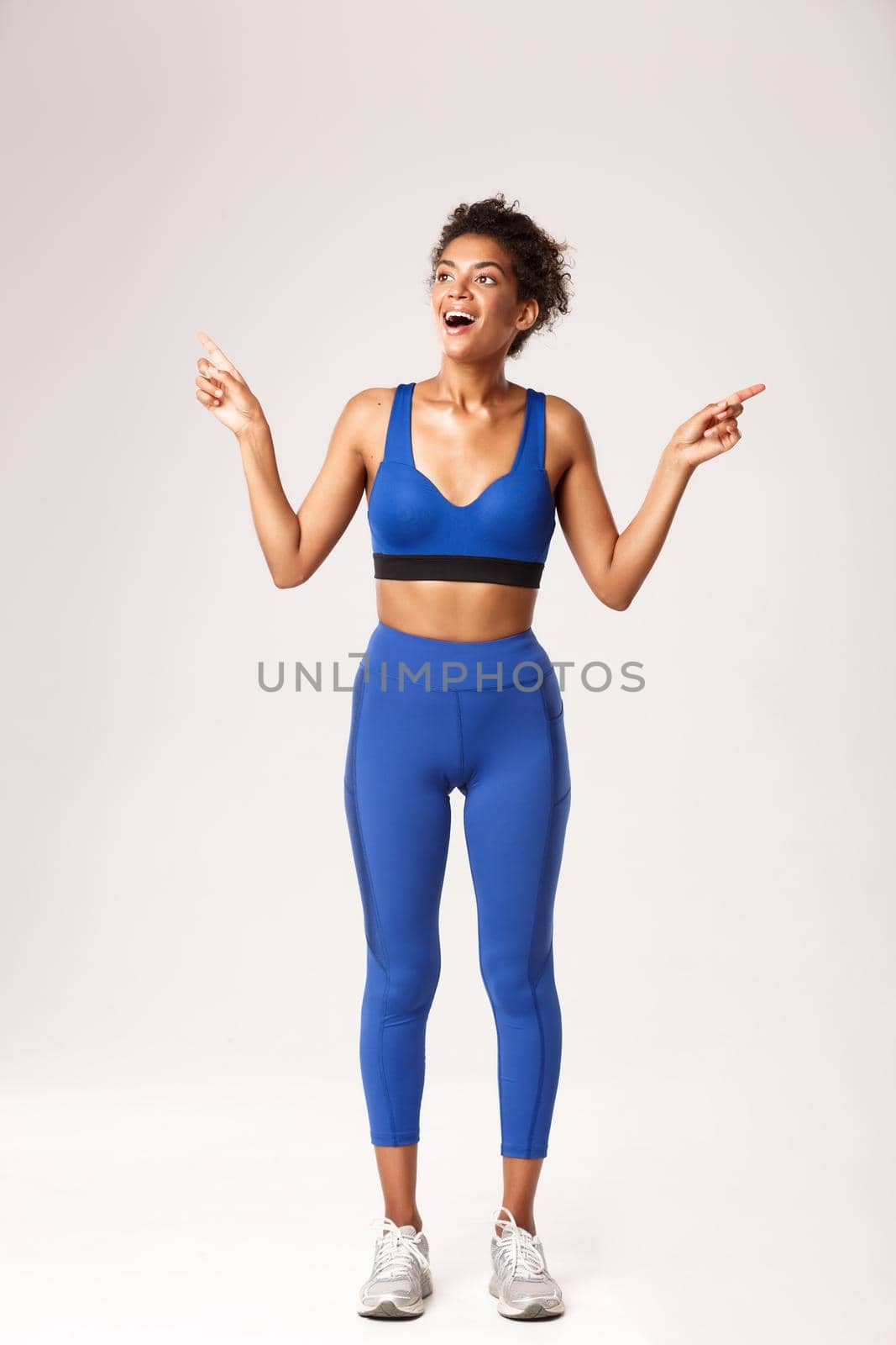 Full length of excited and happy african-american sportswoman, wearing blue sportswear, pointing fingers sideways but looking left with pleased smile, white background.