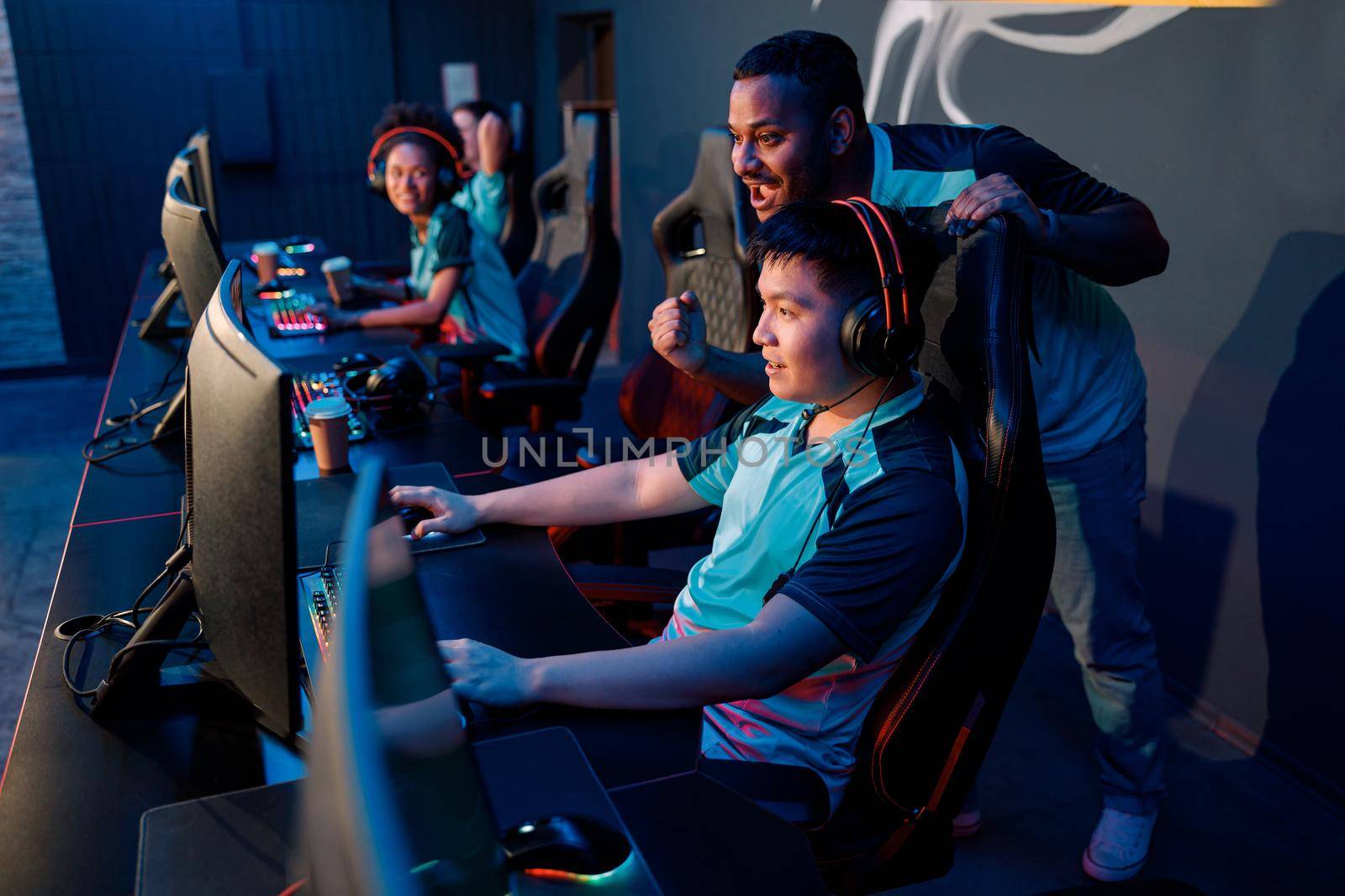 Side view of concentrated Asian male gamer supporting by teammate, winning while playing video games on computer in gaming room