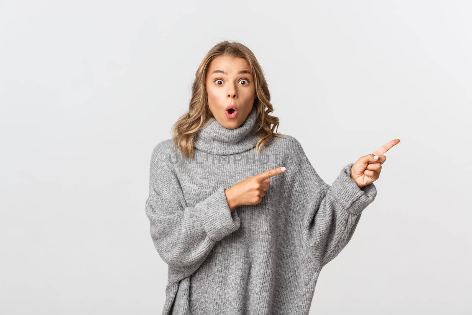 Amazed and excited blond girl in grey sweater showing banner, pointing fingers right and looking at camera, standing over white background.