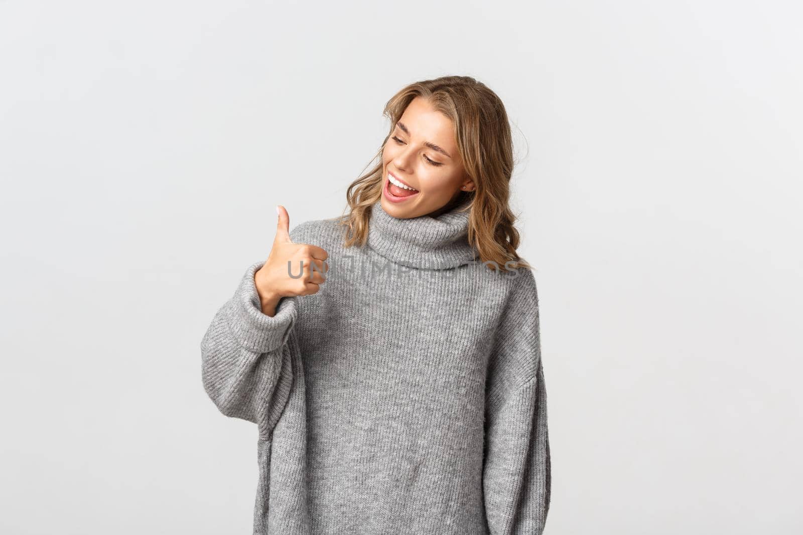 Portrait of cute blond girl in grey sweater, looking at her thumb-up and smiling, standing over white background.