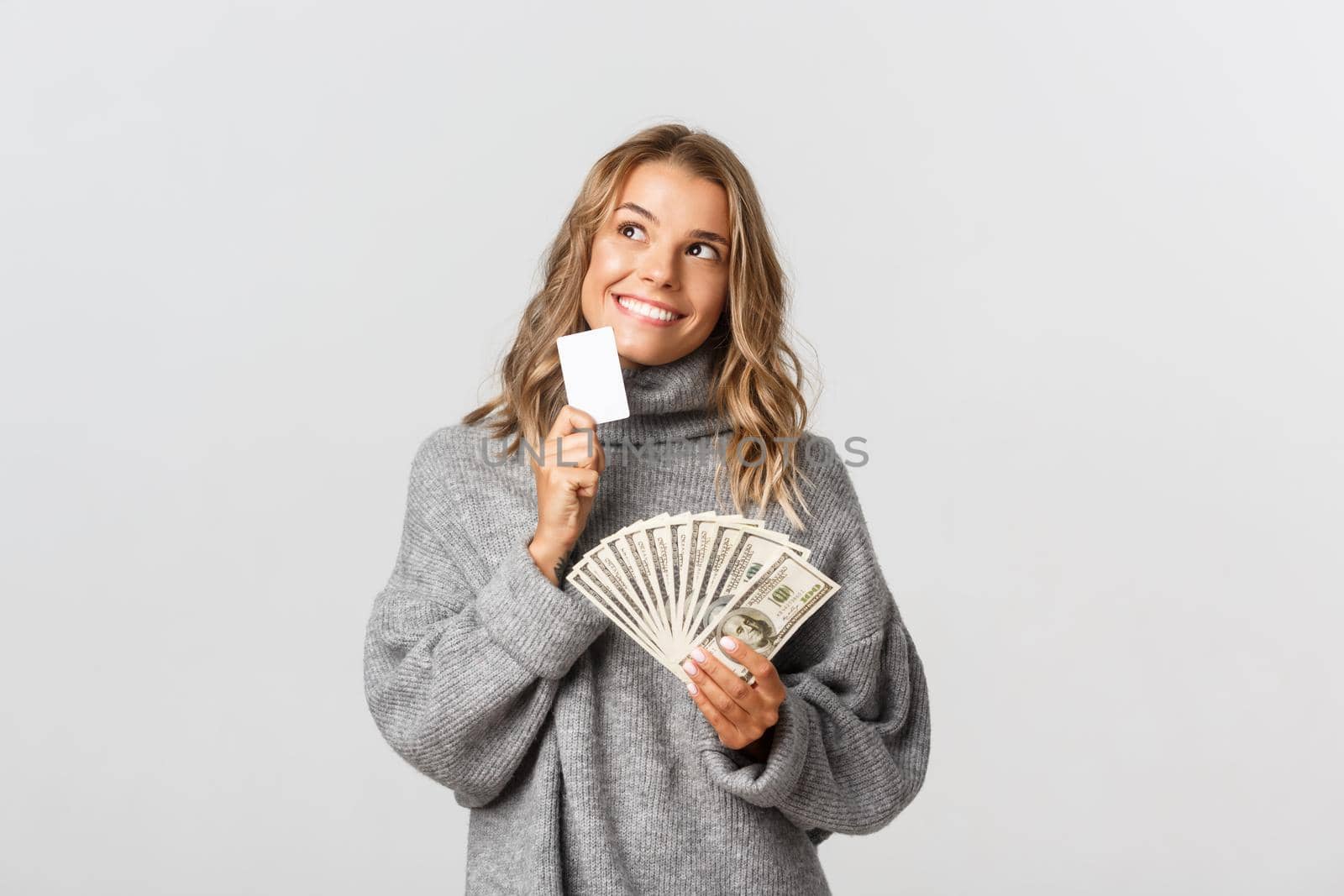 Beautiful blond girl looking thoughtful and smiling, holding credit card and money, standing over white background by Benzoix