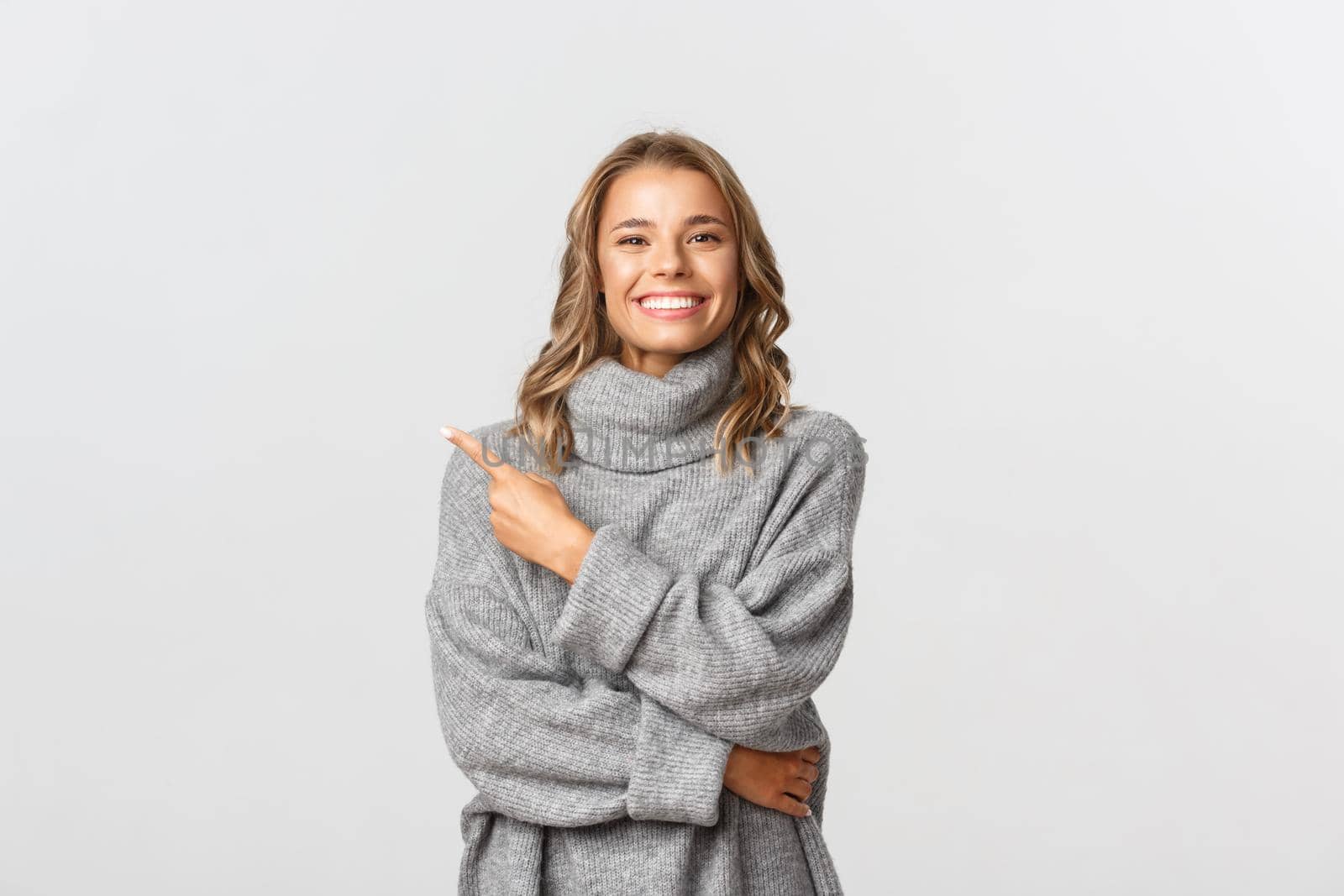 Image of beautiful blond girl in grey sweater pointing finger at upper left corner, showing your logo or banner, standing over white background.