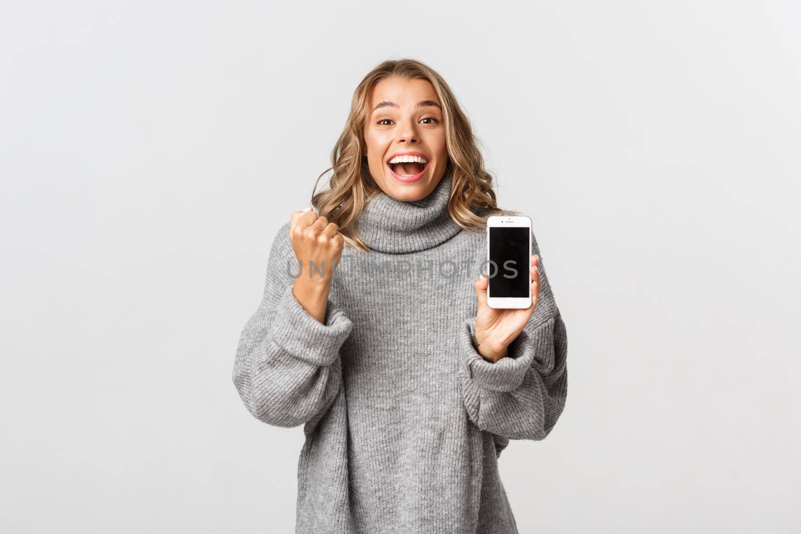 Image of winning happy blond girl, triumphing over online achievement, showing smartphone screen and making fist pump, white background.