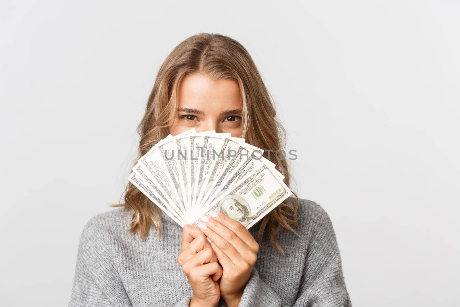 Close-up of excited pretty girl in grey sweater, peeking through money, thinking about shopping, standing over white background.