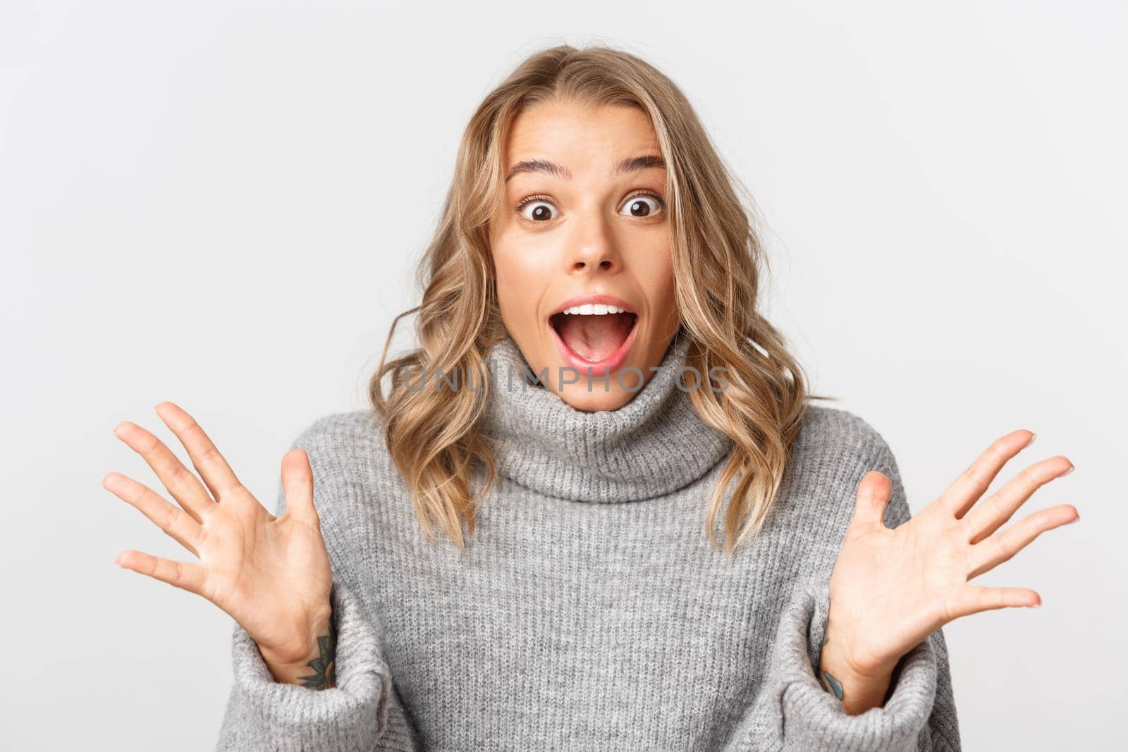 Close-up of beautiful blond girl in grey sweater, screaming for joy, looking surprised, standing over white background.