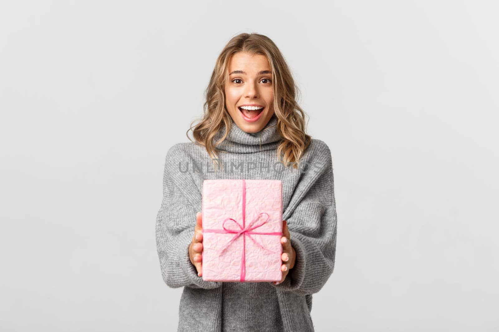Beautiful young woman in grey sweater, giving gift, congratulating someone with birthday, standing over white background.
