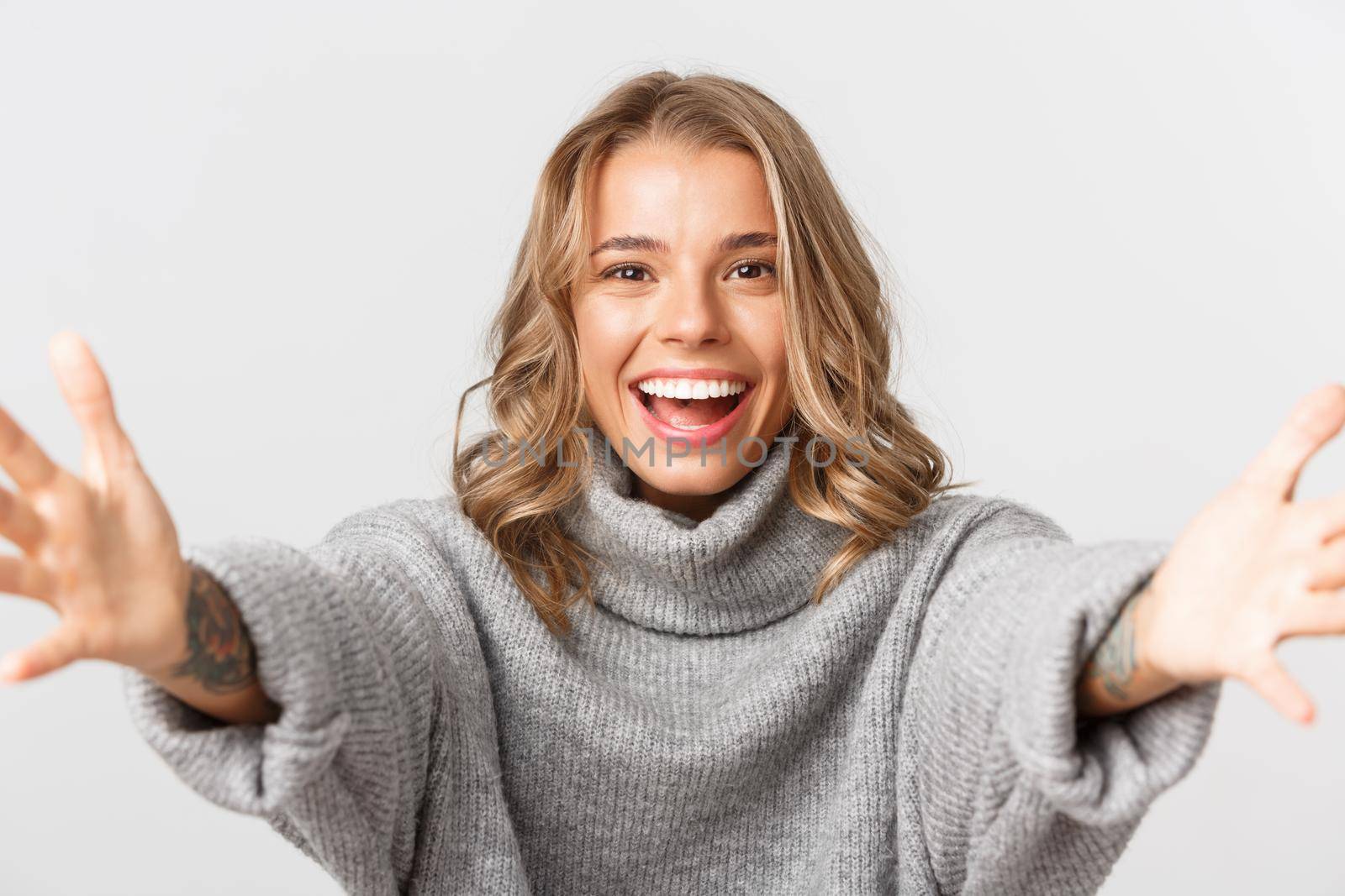 Close-up of happy attractive blond girl smiling, stretching hands forward for hug, reaching for something, standing over white background by Benzoix