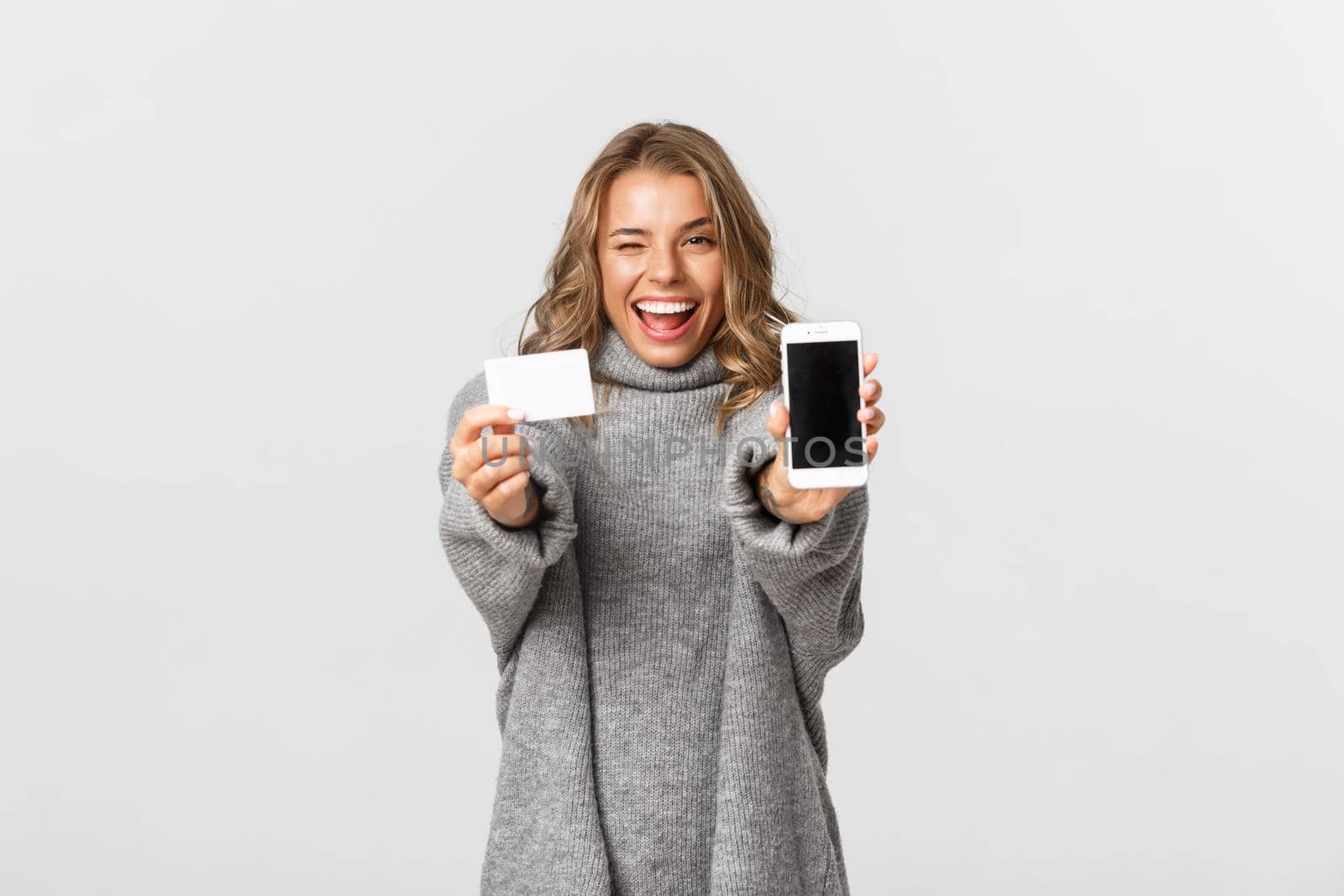 Studio shot of attractive blond girl in grey sweater, showing mobile phone screen and credit card, looking happy, standing over white background.