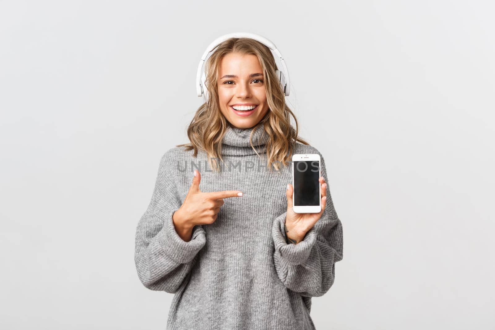 Studio shot of attractive blond girl in grey sweater, listening music in wireless headphones, showing smartphone screen and smiling, white background.