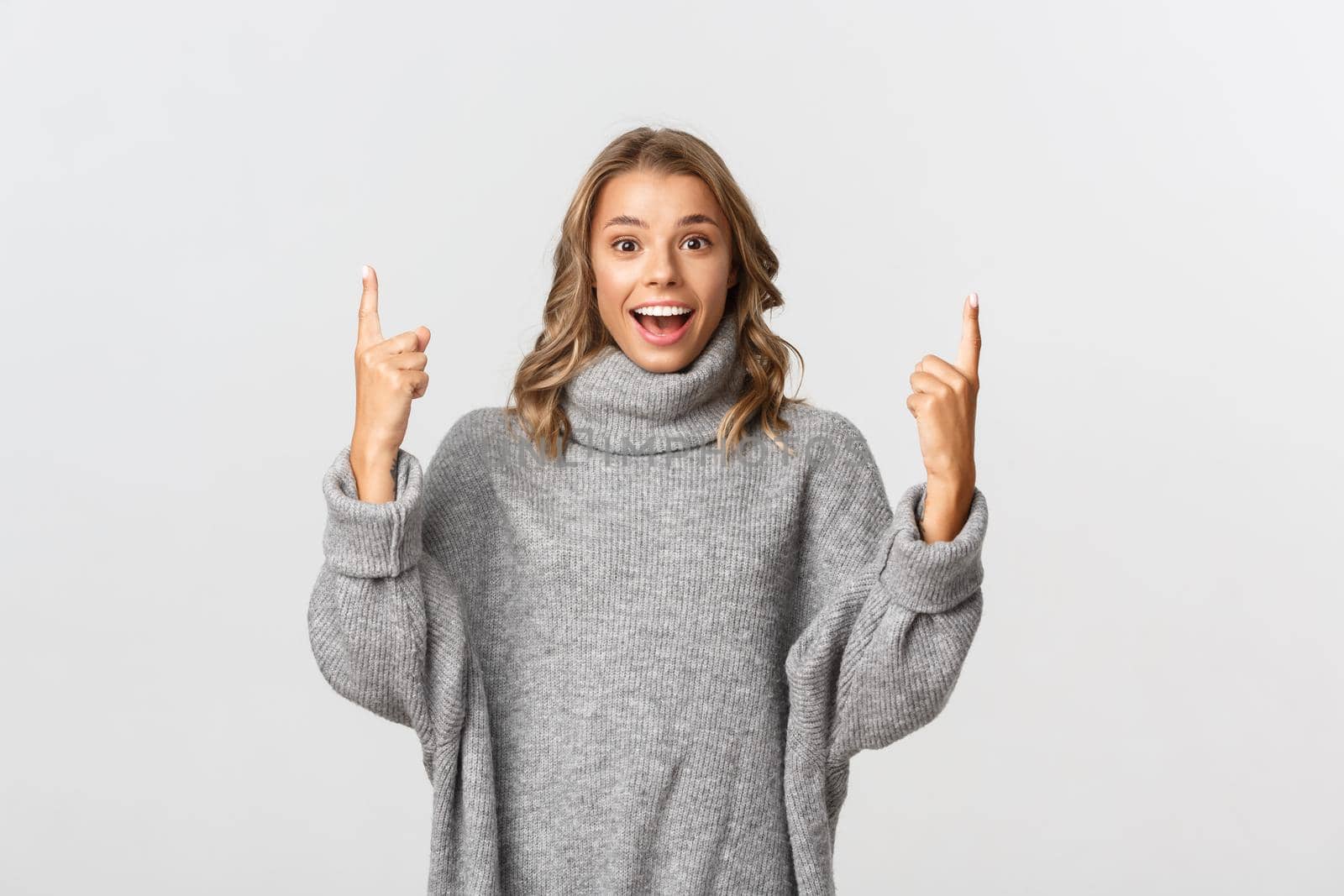Image of excited, smiling blond woman in grey sweater, showing banner, pointing fingers up, standing over white background by Benzoix
