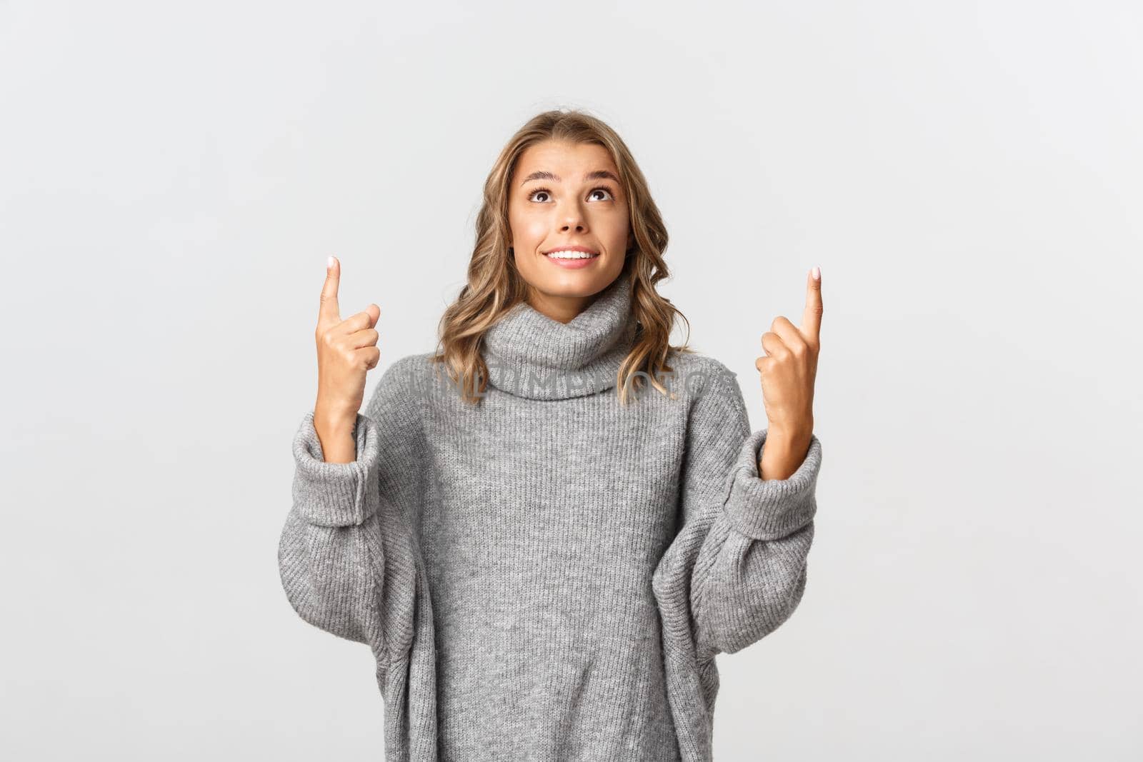 Image of hopeful young woman in grey sweater, pointing and looking up with happy smile, standing over white background by Benzoix