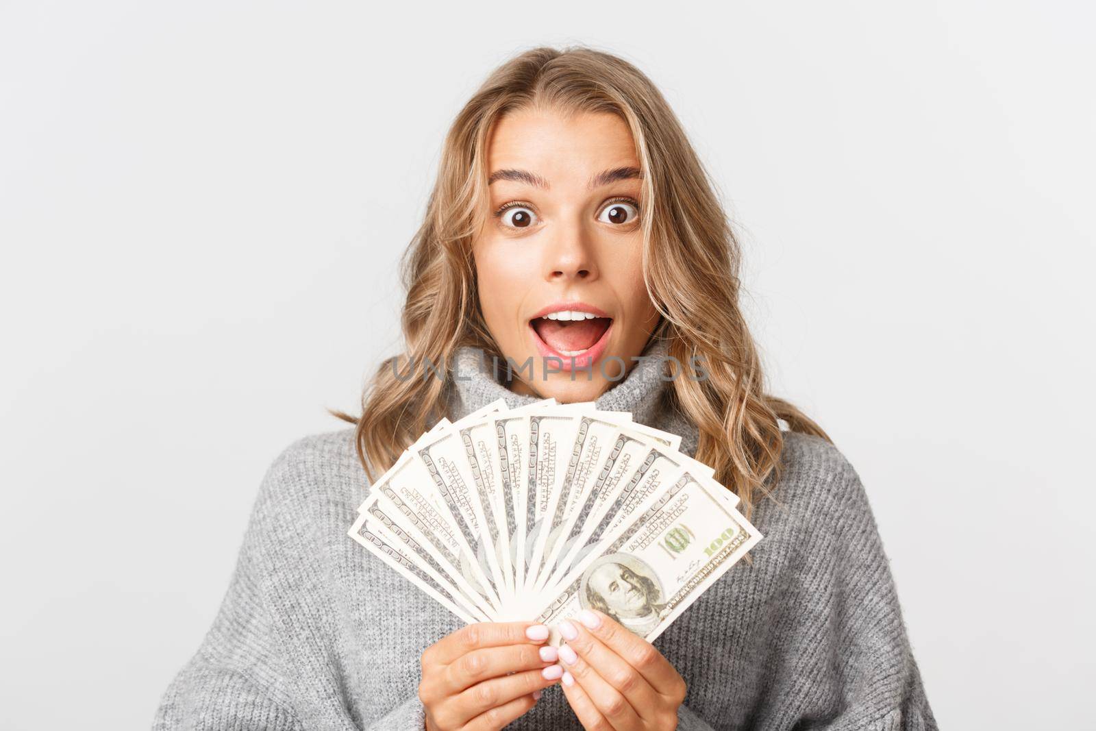 Close-up of excited rich girl with blond short hair, holding money and smiling amazed, standing over white background.
