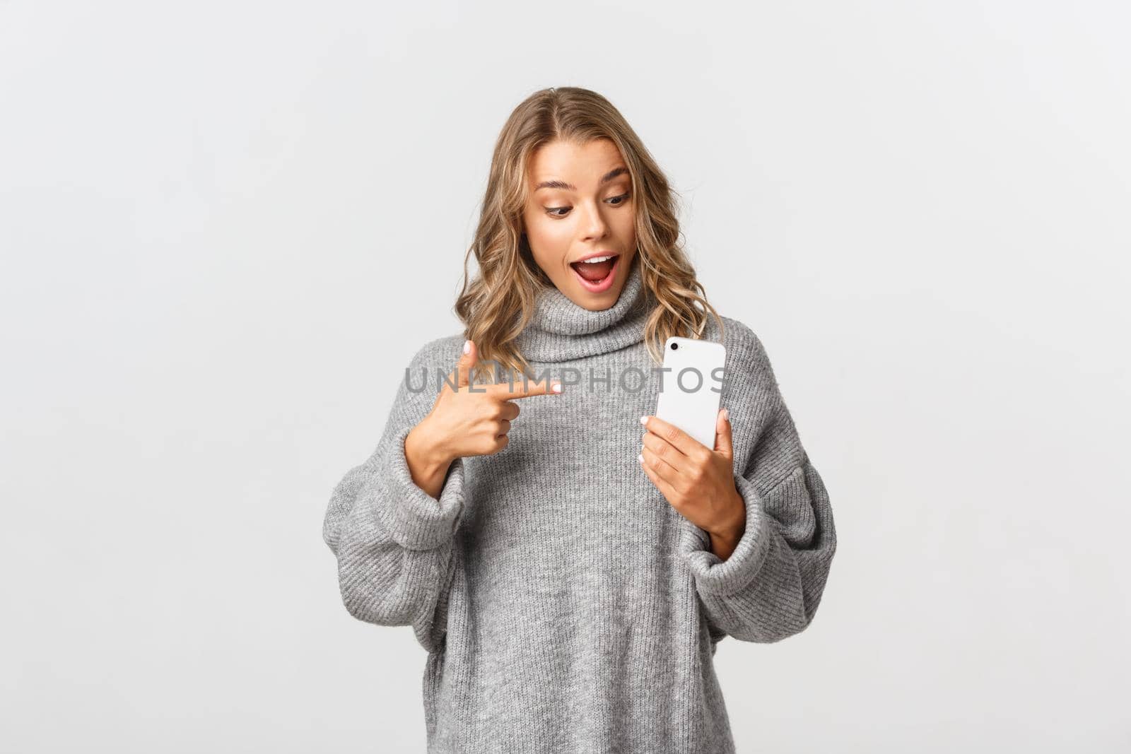 Portrait of attractive caucasian female model in grey sweater, looking amazed and pointing at mobile phone, recommend something, white background.
